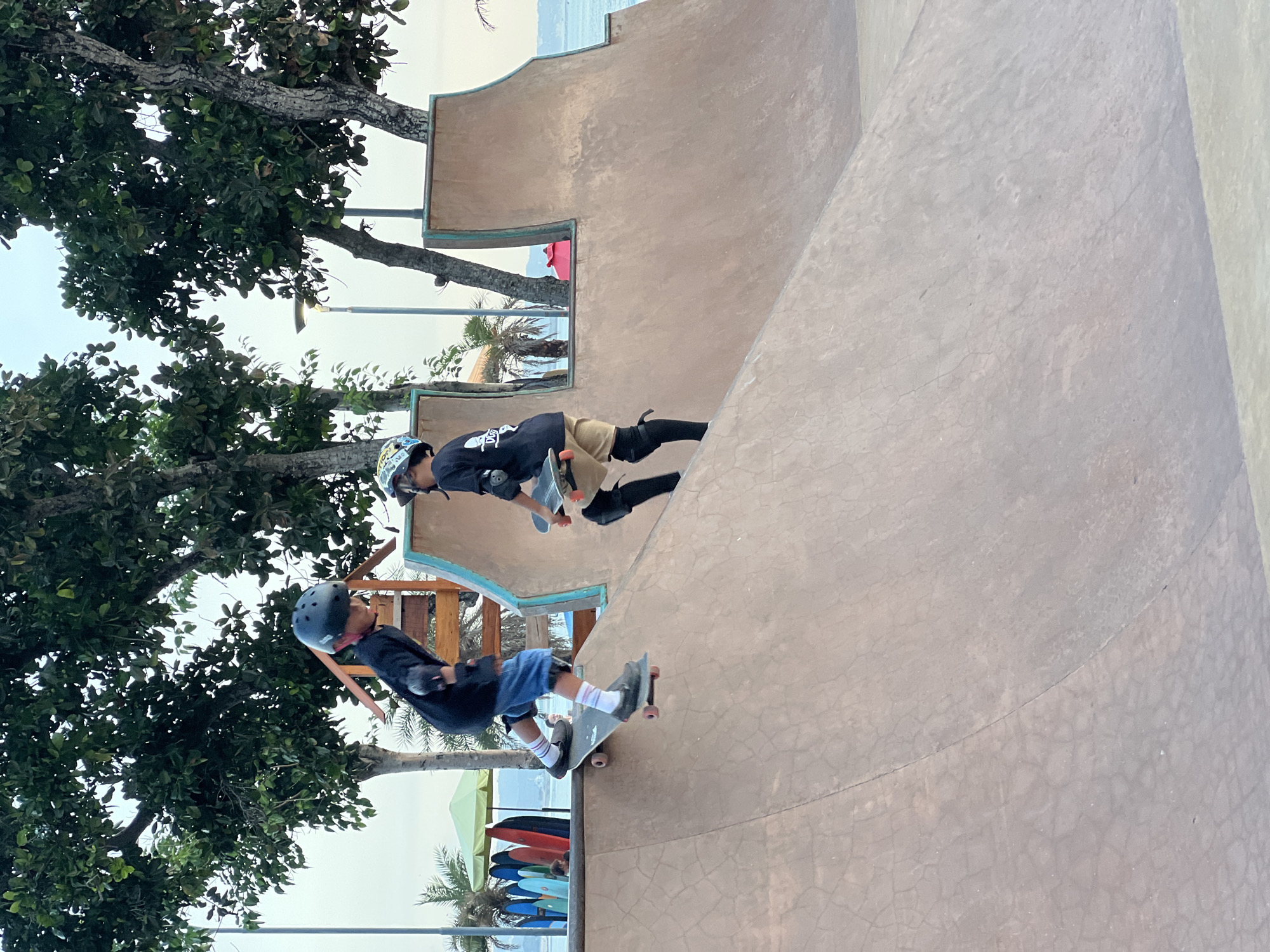 Two young skateboarders wearing protective gear are practicing at a skate park located in Kuta, Indonesia. The skate park features a smooth concrete ramp. Lush tropical trees surround the skate park. Surfboards are stacked nearby.