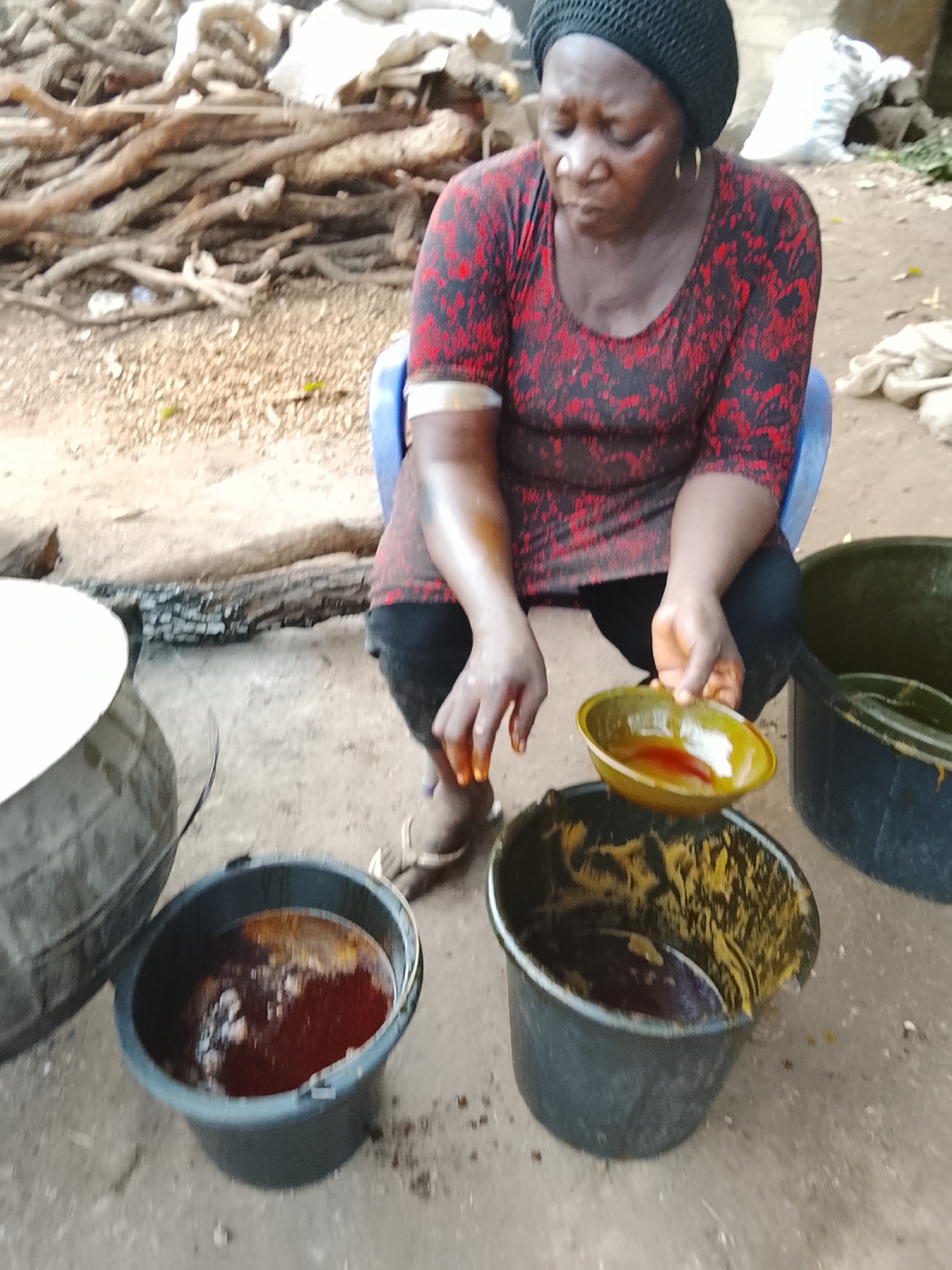 A woman in a black headscarf and red patterned shirt is sitting on a blue plastic chair outdoors. She holds a yellow bowl filled with a red liquid in her right hand and her right hand is stained with the same liquid. Three black plastic buckets are on the ground around her, two containing similar red liquid. A large metal pot with a white lid is to the left of the buckets. In the background, there is a pile of firewood. The location is Unknown, Unknown.