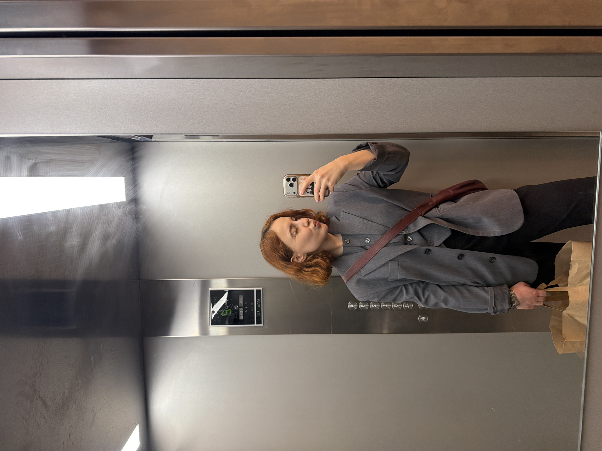 An image taken in Strasbourg, France, captures a mirror selfie of a woman inside what appears to be a modern elevator. The woman, with reddish-brown, medium-length wavy hair, is dressed in a grey blazer over a dark top and black trousers. She wears a dark red crossbody bag and holds a brown paper bag in her left hand. Her right hand is extended, holding a smartphone (an iPhone model with multiple cameras) to take the picture, and she has a slight pout on her lips.
The elevator's interior features polished metallic walls, likely stainless steel, and a large mirror that reflects the woman and parts of the cabin. Horizontal metallic bands are visible at the top of the elevator. To the woman's left, an elevator control panel is embedded in the wall. It features a digital display showing "03:41" and "C8:00". Below the display, a vertical row of circular buttons for floor selection is visible, with numbers ranging from "4" to "15," along with standard door open and close symbols. Bright, rectangular light sources are reflected prominently in the mirror, and some streaks or smudges are visible on the mirror's surface. The setting appears to be indoors, likely during the day or evening, given the artificial lighting.
