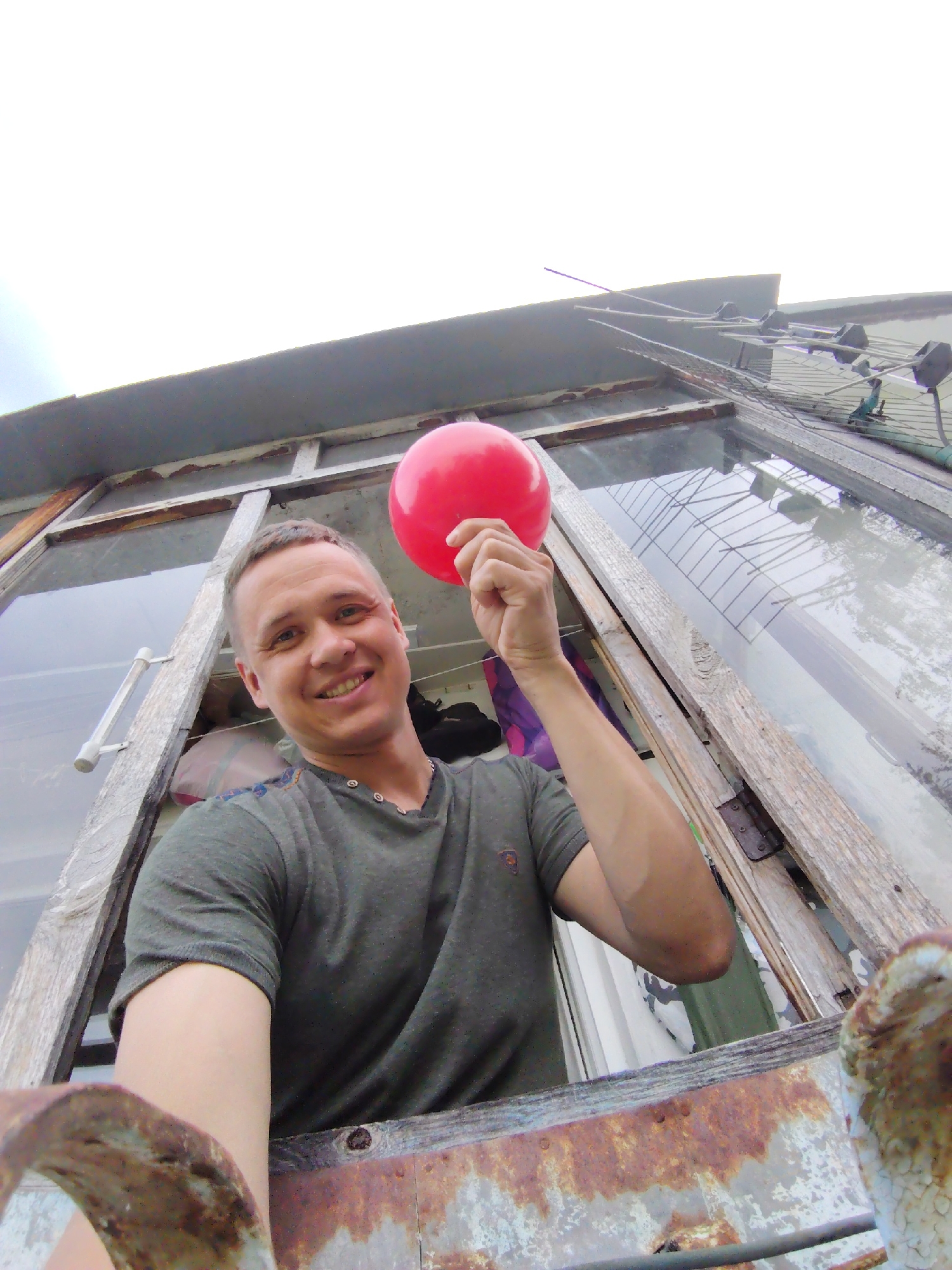 A man is holding a red balloon while looking out of a window. The window frame is old and appears to be located on an upper story. The exterior appears weathered, with rust visible on the lower frame. An external television antenna is visible above the man and window. A location in the city of Chaikovsky, Russia is specified.