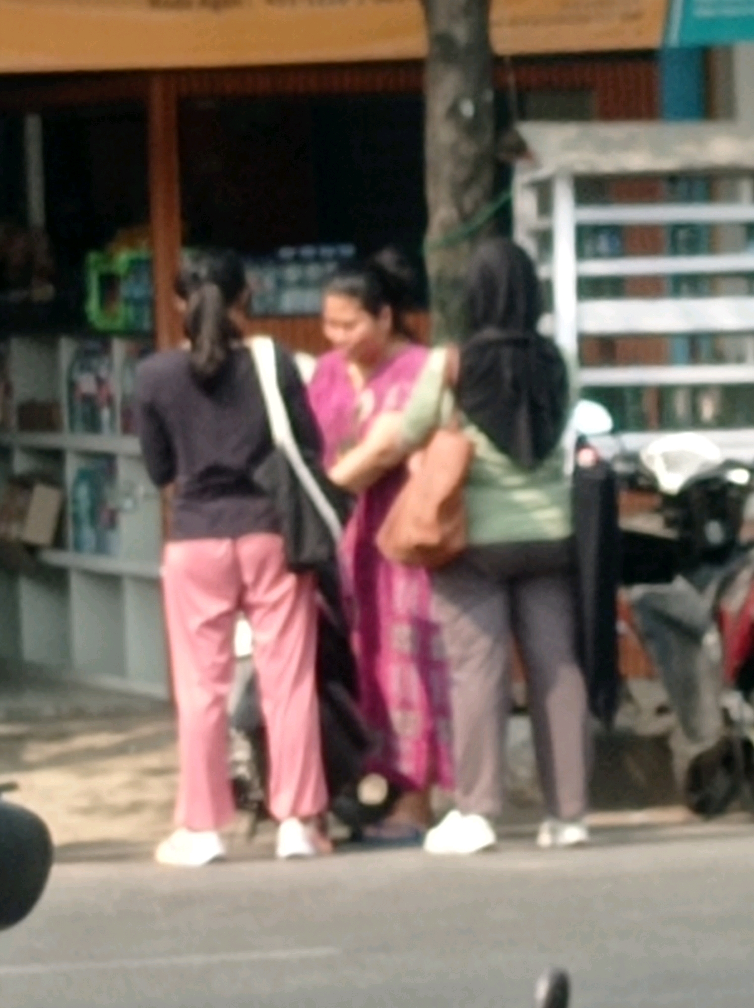 The image shows three individuals standing on a paved surface, possibly a street or sidewalk, in front of a building with shelves stocked with items. The building appears to be a shop or market. The first person on the left is wearing a dark top and pink pants, with a dark bag slung over their shoulder. They are facing away from the camera. The second person, in the middle, is wearing a bright pink patterned dress and is facing towards the left. They have a brown bag slung over their shoulder. The third person on the right is wearing a black hijab and a green top, with a dark bag or backpack. They are facing away from the camera. In the background, there are shelves filled with various objects and a motorcycle is visible to the right. The overall scene suggests a bustling street in Tuban, Indonesia.
