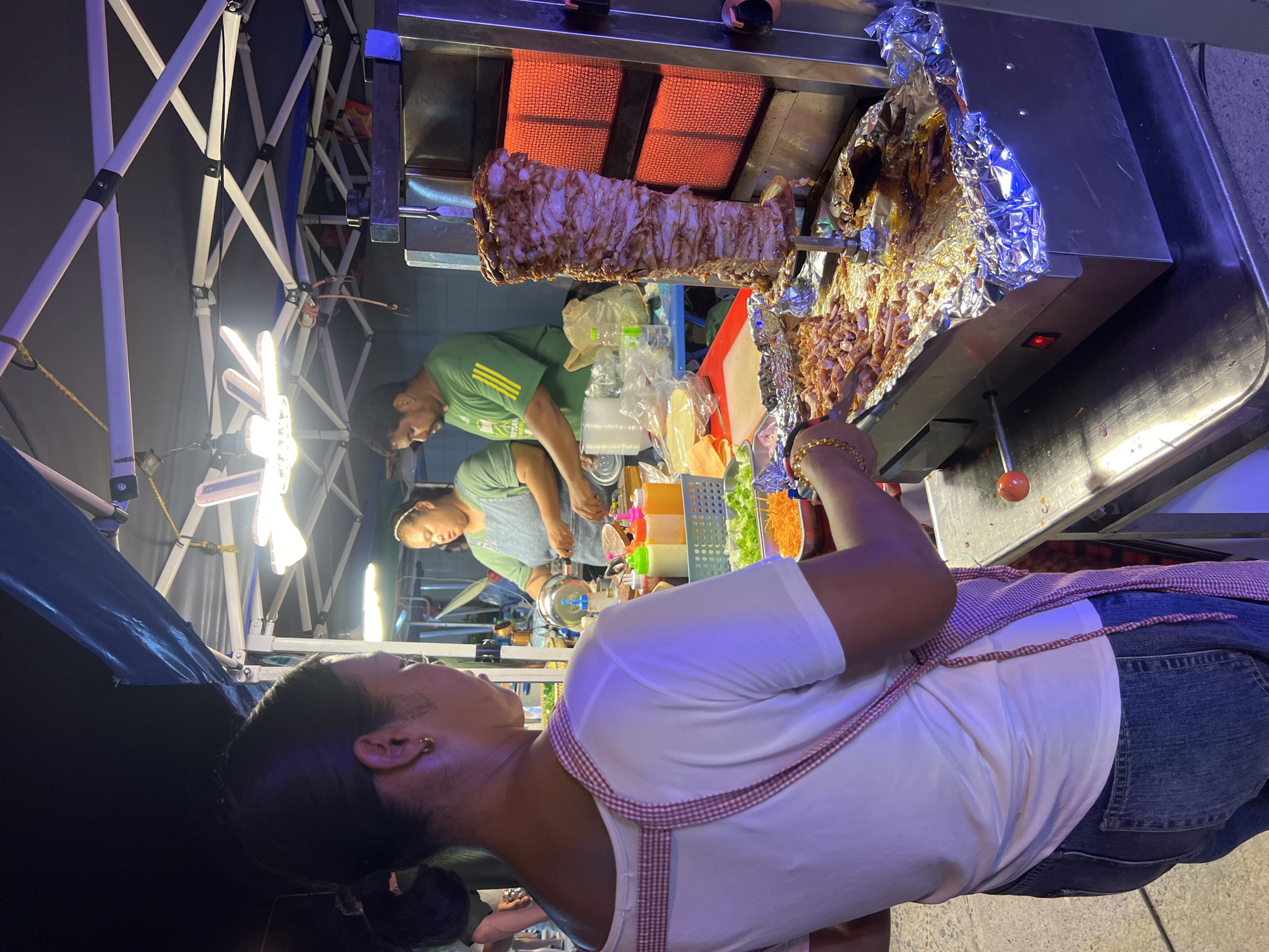 A street food stall in Ko Pha-ngan District, Thailand, is shown at night. A large vertical rotisserie with seasoned meat is being cooked, and shaved meat is being prepared on a metal tray below. A woman in a white shirt and jeans, wearing a checkered apron, is standing to the left, holding a spatula. Behind her, two people are visible working at the stall. One person, wearing a green shirt, is standing and appears to be preparing food, while another person, wearing a blue apron, is behind a counter. The stall is set up under a tent structure with bright overhead lighting. Various condiments and fresh ingredients like shredded carrots and lettuce are arranged on the counter. The overall scene depicts a busy food vendor operating at night.