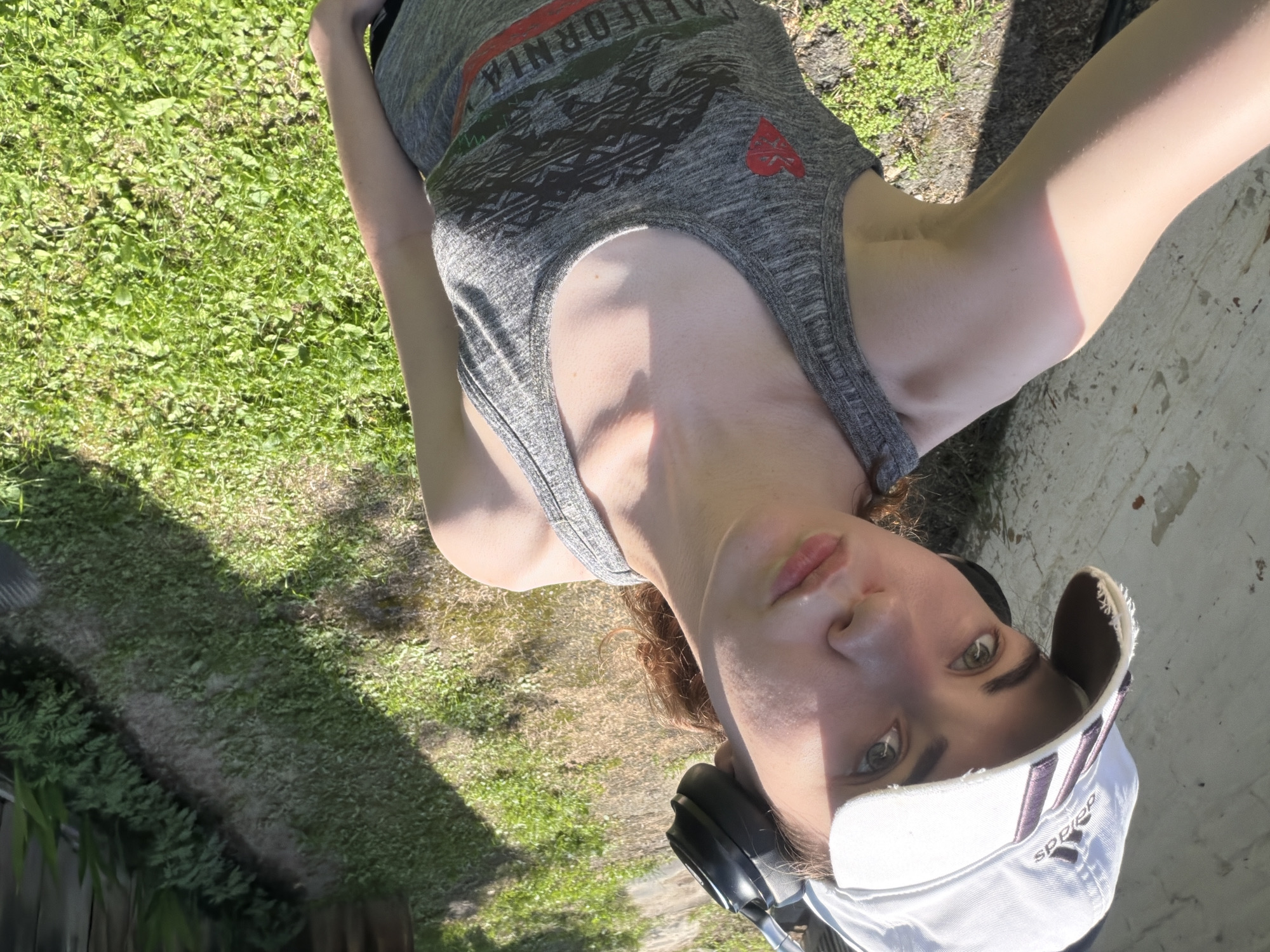 The media file is a portrait-style selfie of a young person wearing a grey tank top and a white baseball cap. The subject has reddish-brown hair and is looking directly at the camera with a neutral expression. They are wearing headphones around their neck. The background consists of green grass and a concrete surface. The lighting suggests it is daytime, with sunlight casting shadows. Visible text on the tank top reads "CALIFORNIA" in red and green letters, with a graphic below it and a red heart shape. The baseball cap has the Adidas logo visible. The scene appears to be outdoors, possibly in a backyard or park. There are no clear indications of a specific event taking place, beyond the act of taking a selfie.