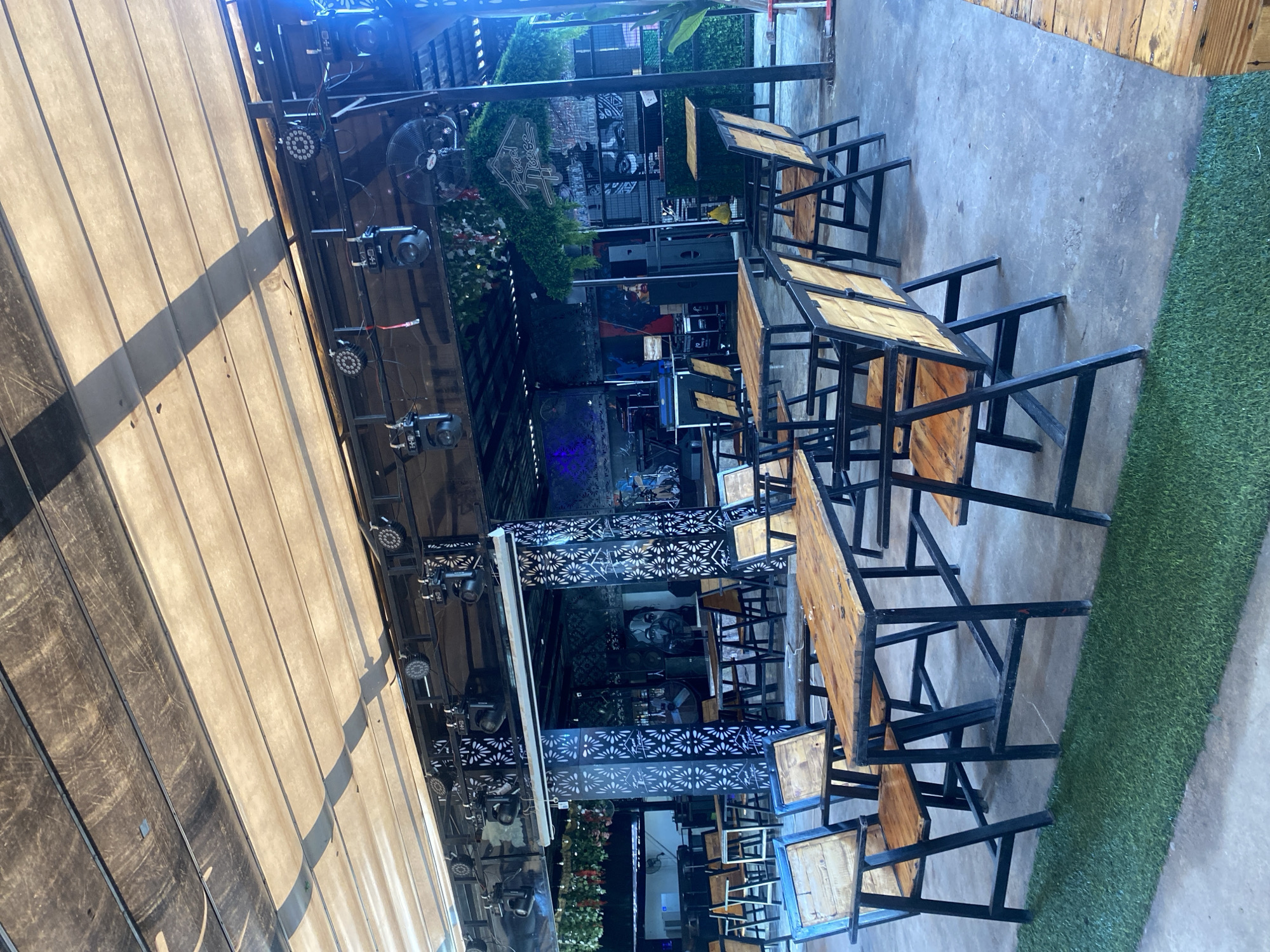 The image displays an unoccupied, semi-open-air entertainment venue, likely a bar or live music establishment, captured during daylight hours.
**Environment:** The overhead structure features a retractable, light-colored fabric awning, showing signs of wear and soiling, partially covered by darker fixed panels. A robust black metal truss system supports numerous stage lighting fixtures, including several moving head lights (some bearing "HH D" markings) and LED par cans, alongside at least one industrial-style ceiling fan.
**Furnishings:** The main seating area consists of multiple rectangular wooden tables with dark metal frames, surrounded by matching chairs. These chairs feature wooden slatted seats and backs, supported by dark metal frames; a chair in the left foreground exhibits blue accents on its frame. All tables and chairs are currently empty and arranged for patronage.
**Background/Stage Area:** Towards the rear of the venue, a stage area is visible, equipped with musical instruments such as a keyboard and a drum set, flanked by large sound speakers. Structural pillars are decorated with a white patterned design against a dark background. Wall murals, including a partially visible stylized face, adorn the dark rear walls. Artificial green foliage hangs from overhead beams and decorates a sign that reads "Road House."
**Floor:** The floor is primarily grey concrete, exhibiting varied tones and some marks. A strip of artificial green turf runs along the bottom foreground of the image, separating the main seating area from an unseen adjacent space.
**People/Activity:** There are no individuals present in the scene, and no discernible activity is taking place, suggesting the venue is either closed or awaiting guests.
**Location:** The image itself does not contain any visual identifiers to confirm its location in the city of Howayi, Nigeria.