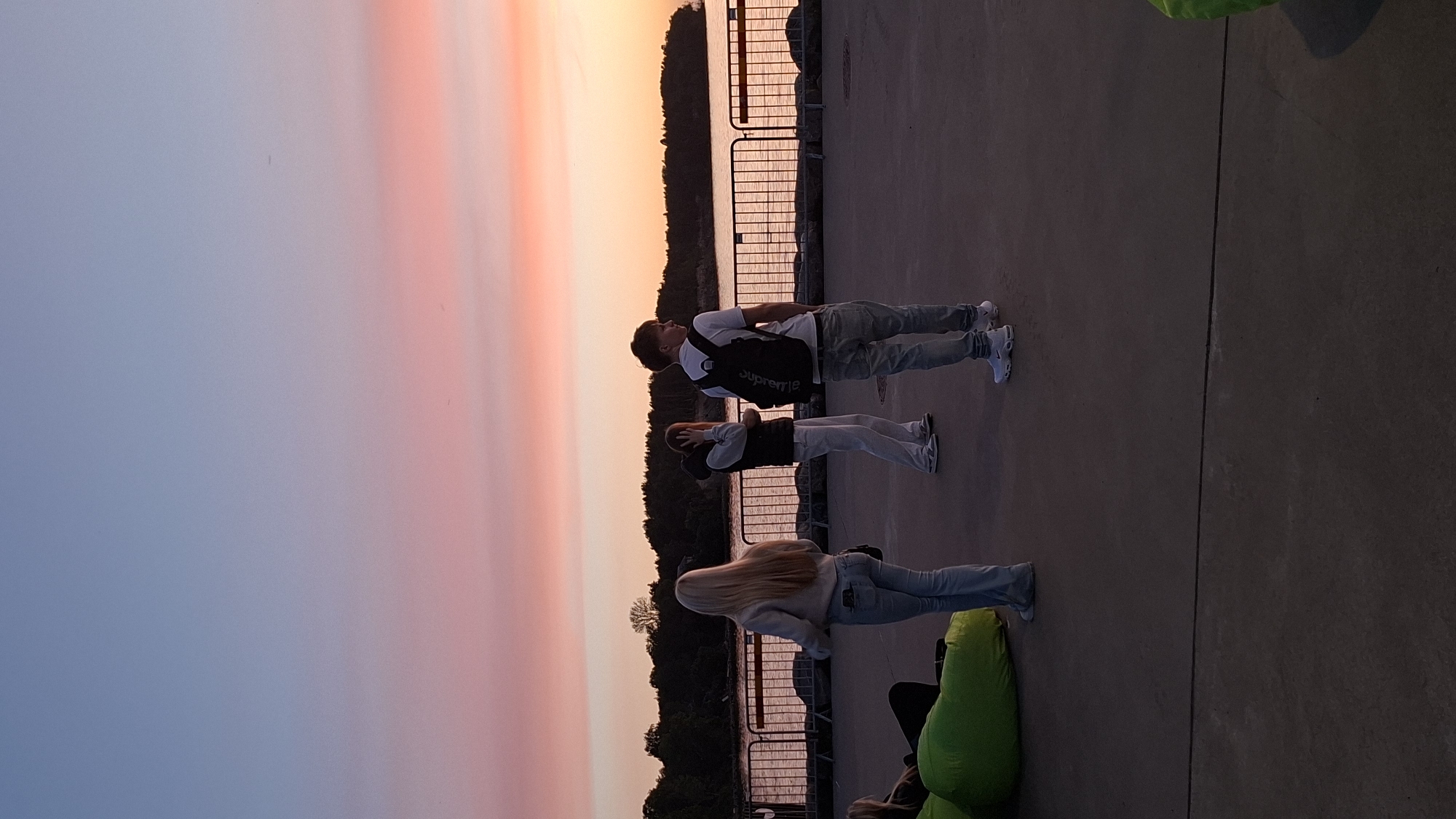 In Unknown, Unknown, at sunset, three young individuals are seen standing on a paved area facing a body of water. The sky displays a gradient of blue transitioning into pink and orange hues. A lush, tree-lined shore is visible across the water, with a metal railing running along the edge of the paved area. The person on the left is a woman with long blonde hair, wearing a light-colored hoodie and blue jeans. To her right, a person with dark hair is wearing a dark vest over a hooded sweatshirt and white pants, with their hands near their head. The third individual, a young man with reddish-brown hair, is on the far right, wearing a white t-shirt, blue jeans, and a black backpack. They are all facing away from the camera. In the foreground, a large, bright green beanbag chair is partially visible on the left. The ground is a dark, textured concrete surface.