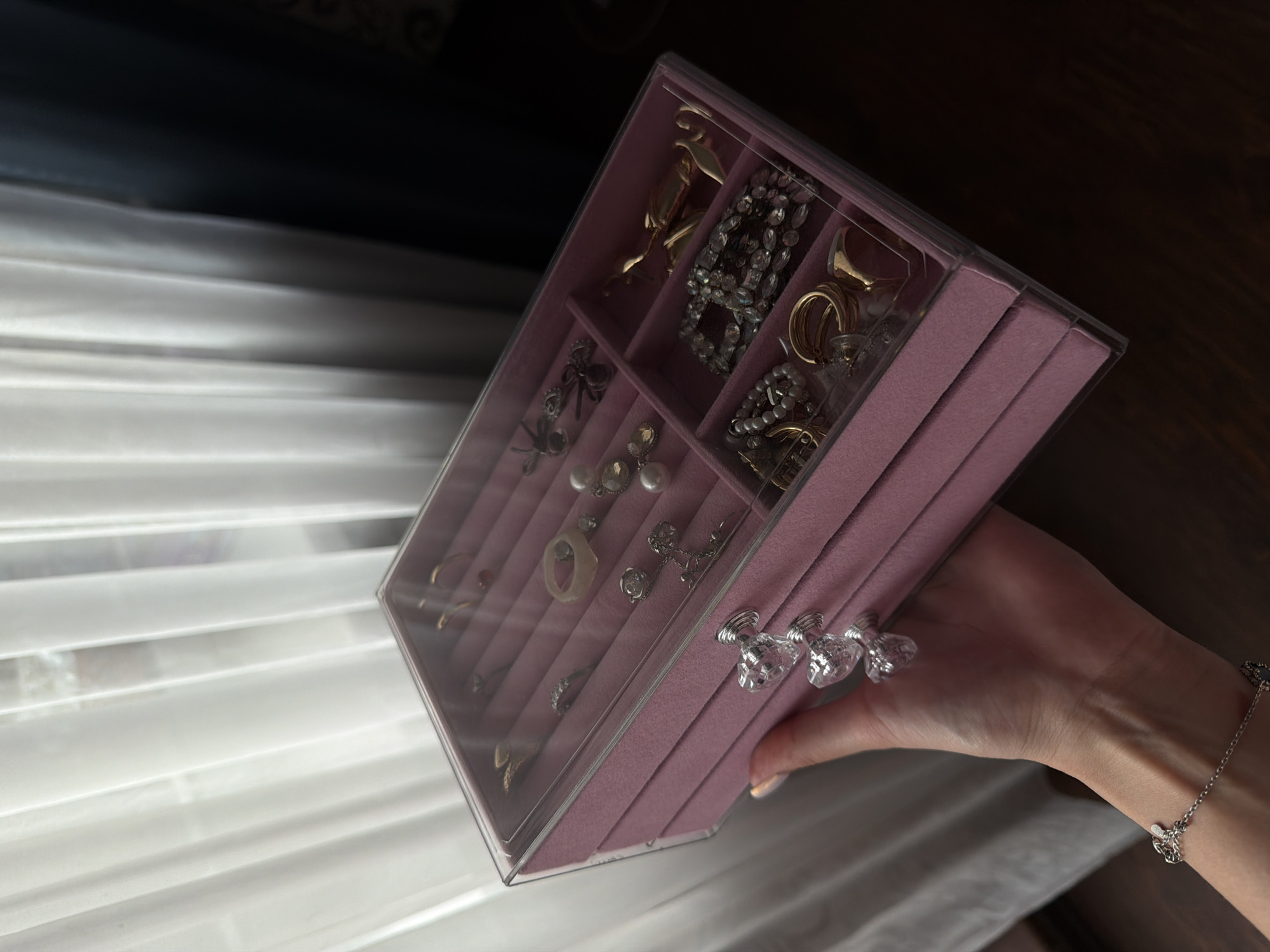 A person's hand holds a pink, multi-tiered jewelry box filled with various pieces of jewelry. The jewelry box has a clear lid and crystal-like drawer pulls. The jewelry inside includes rings, necklaces, bracelets, and earrings. The background consists of white sheer curtains and a dark, wooden surface.