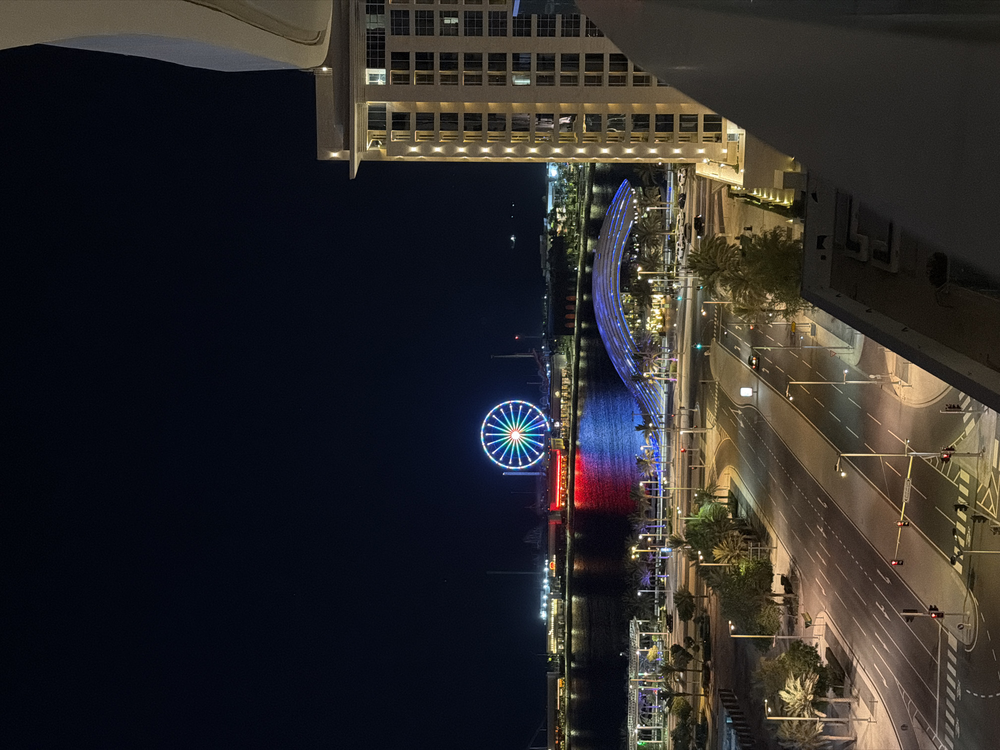 The image presents a vibrant night-time view of a cityscape in Lusail, Qatar, captured from an elevated perspective, likely a balcony or high floor of a building. The scene is dominated by a large, colorfully lit Ferris wheel situated on the far side of a body of water, its spokes displaying a rainbow of red, green, blue, yellow, and orange lights.
Running parallel to the water is a multi-lane road or highway, clearly defined by bright streetlights and road markings. Traffic lights are visible at intersections, indicating active but not congested traffic flow. Adjacent to the road and along the waterfront, palm trees and other landscaping are illuminated by ground-level lights.
A prominent, arching structure, possibly a pedestrian bridge or a decorative element, spans across the water, glowing with intense blue lights. The body of water itself reflects the numerous artificial lights from the surroundings, creating shimmering streaks of color, including a distinct red reflection near the base of the Ferris wheel. In the distance, faint lights from what appear to be boats or distant structures dot the horizon over the water.
To the right of the vantage point, modern multi-story buildings with illuminated windows and balconies create a backdrop for the scene. The sky is dark and clear, characteristic of a cloudless night. No individuals are distinctly visible, and no specific event is taking place, but the scene portrays a lively and well-developed urban environment after dusk, showcasing modern architecture and intricate lighting. There is no visible text in the image.