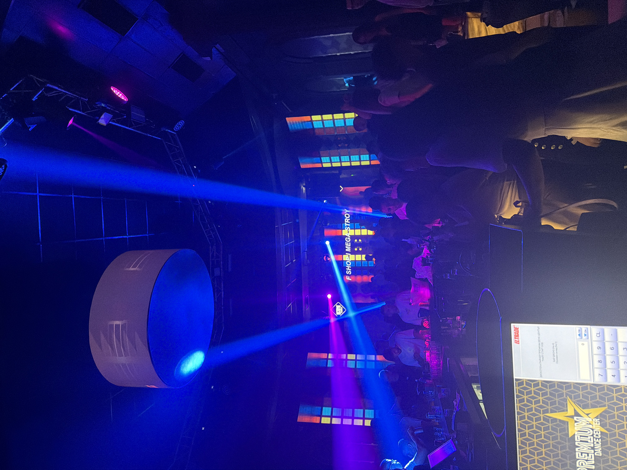 The image depicts the interior of a nightclub or dance center, bathed in vibrant blue and purple stage lighting. A large, cylindrical light fixture hangs from the ceiling, illuminating the space. Beams of light cut through the darkness, and colorful, rectangular panels are visible on the back wall. A crowd of people is gathered, some standing and others appearing to be at a bar or counter. A portion of a digital display with a keypad and the logo "PREMIUM DANCE CENTER" is visible in the foreground. The overall atmosphere suggests a lively event or party. The image appears to have been taken within the city of Veliko Tarnovo, Bulgaria.