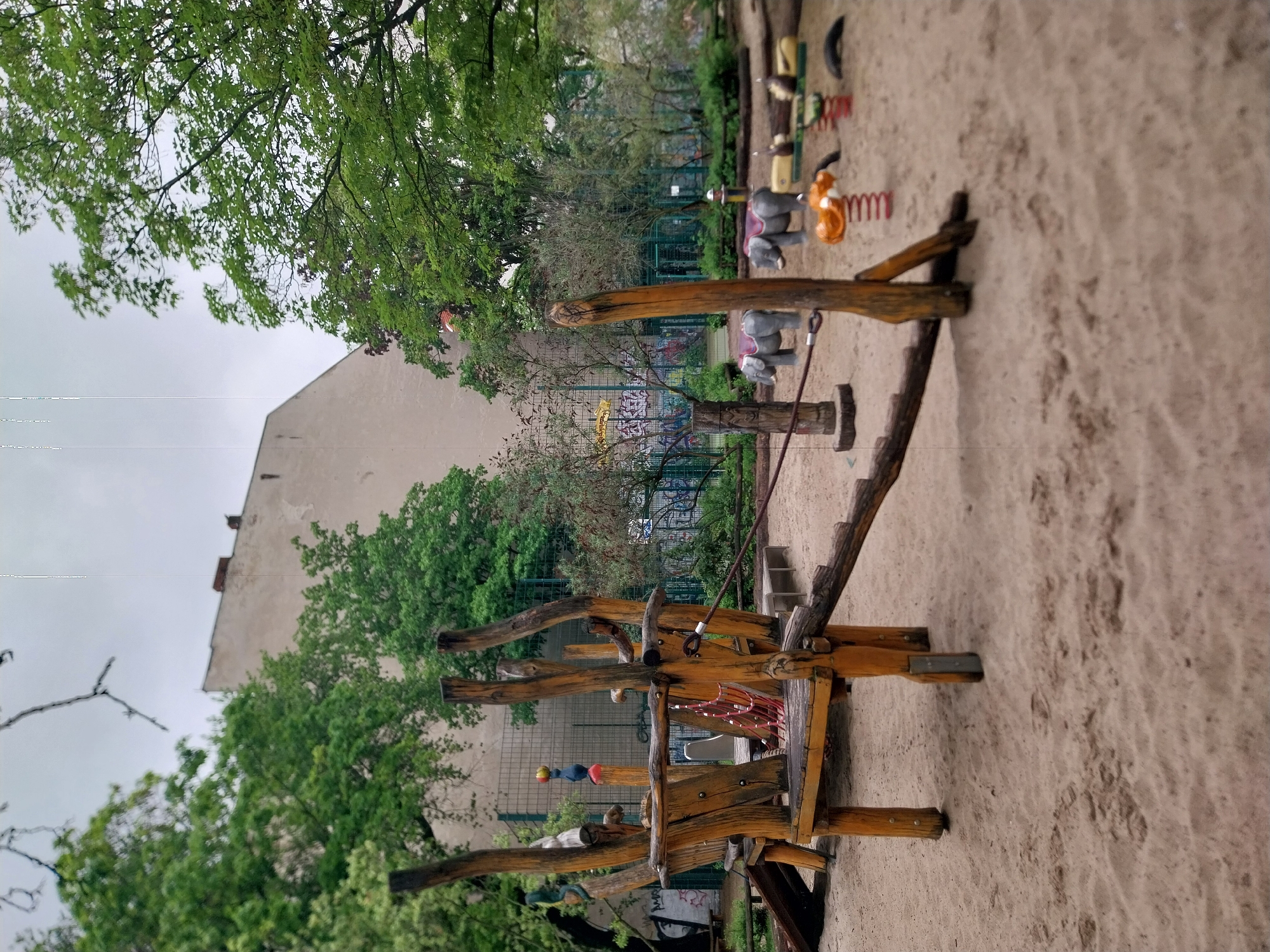 The image shows a playground in Berlin, Germany. The playground is sandy and includes a wooden play structure with climbing elements. In the background, there is a building and a fence with graffiti. Other playground equipment, including spring riders shaped like animals, is visible. Green trees and foliage surround the playground. The sky is overcast.