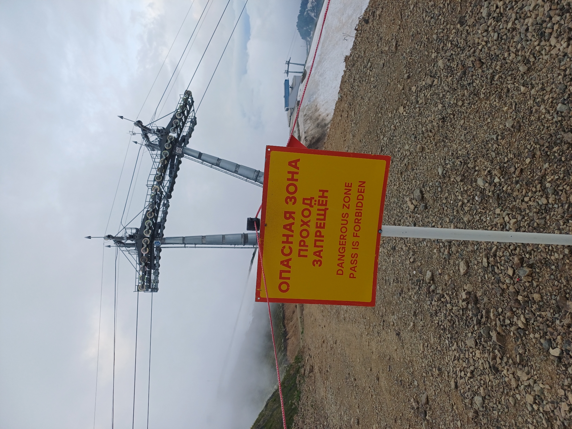 The image shows a yellow sign with red trim, which reads "ОПАСНАЯ ЗОНА ПРОХОД ЗАПРЕЩЁН" in Russian and "DANGEROUS ZONE PASS IS FORBIDDEN" in English. The sign is positioned in a rocky, mountainous area, likely a ski resort in Estosadok, Russia, given the presence of ski lift infrastructure in the background. The ski lift towers and cables are visible against a cloudy sky. A red rope barrier is set up across the path in front of the sign, further reinforcing the warning. The ground is covered in gravel and some patches of snow can be seen on the slopes in the distance. The overall environment appears to be an outdoor, mountainous terrain with evidence of winter sports infrastructure. There are no people visible in the image.