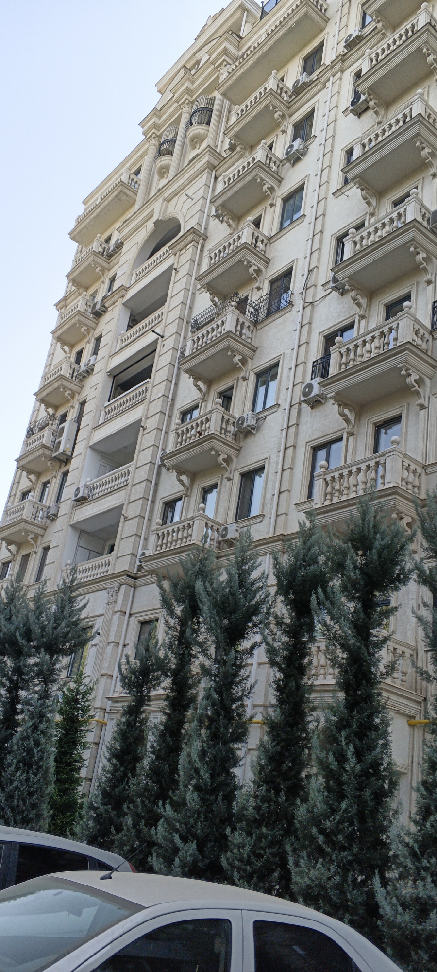 The image shows a tall, ornate apartment building with a light-colored facade and multiple balconies. The building has classical architectural elements such as columns and decorative carvings. Several air conditioning units are visible on the balconies. In the foreground, a line of tall, slender evergreen trees partially obscures the lower part of the building. The sky is clear and light. The building is likely located in Tashkent, Uzbekistan, a city known for its blend of Soviet-era and modern architecture, with many new apartment complexes featuring decorative facades and balconies.