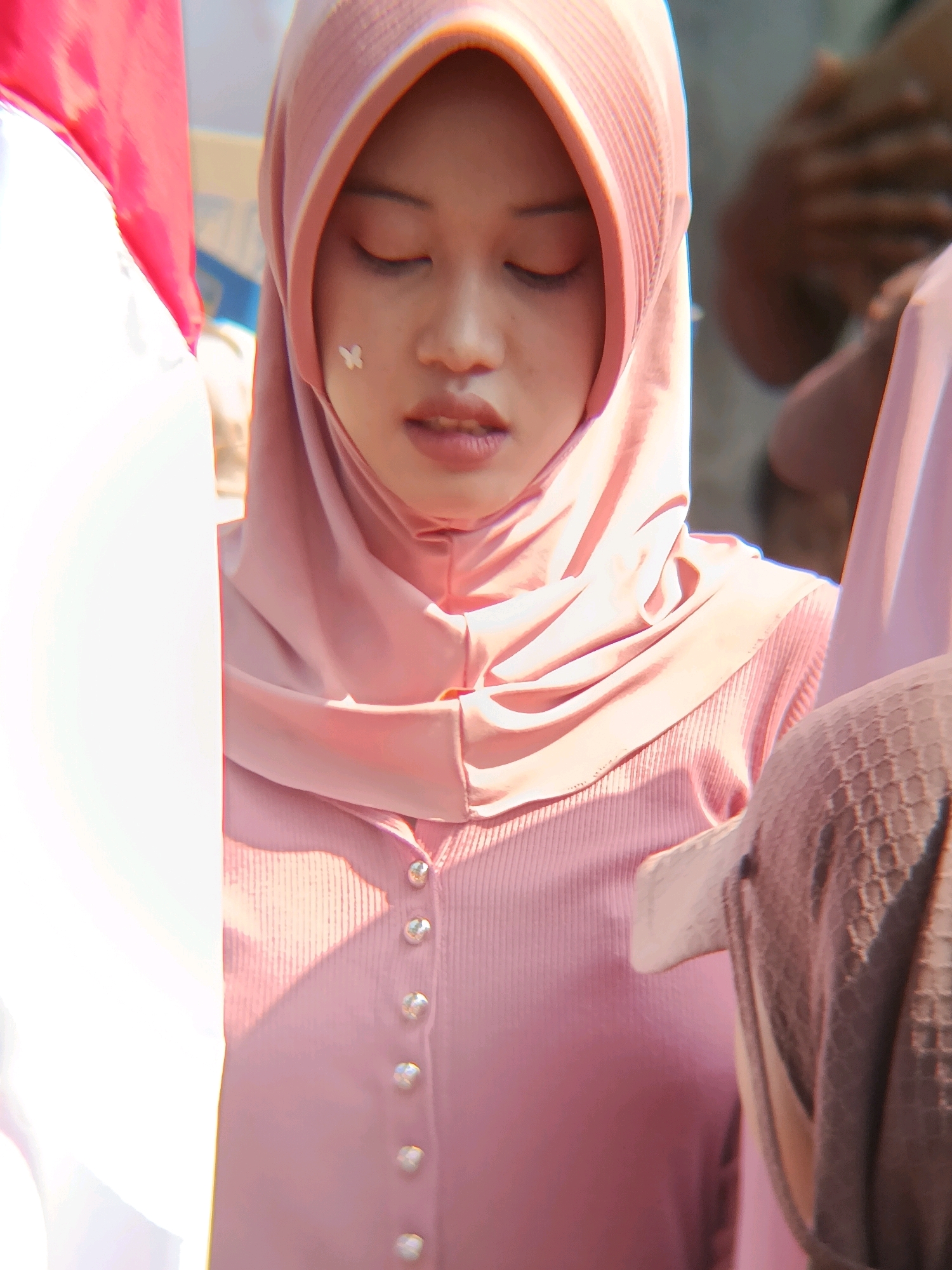 The image is a close-up portrait of a young woman wearing a pink hijab and a pink ribbed top with pearl-like buttons. She has her eyes closed and a small, light-colored butterfly sticker on her cheek. To her left, a portion of the Indonesian flag, red and white, is visible. In the background, blurred figures and a part of a grey patterned garment suggest an outdoor setting, possibly a public event in Binangun, Indonesia, given the flag. The lighting is bright, creating highlights and shadows on the fabric of her clothing.