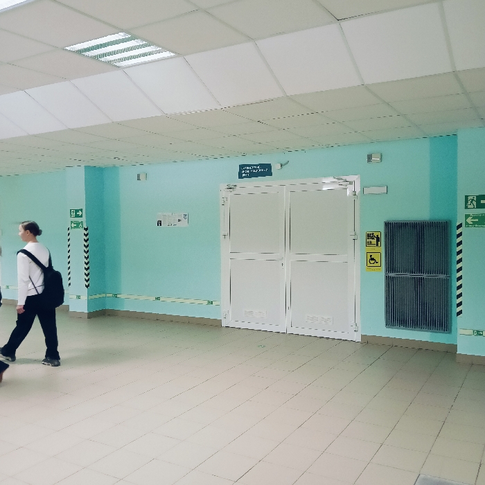 The image displays an interior corridor within an educational institution, specifically identifiable as a learning environment by prominent Cyrillic signage, located in Chaikovsky, Russia. The walls are painted a uniform light blue (cyan), and the floor consists of light-colored square ceramic tiles. A white modular drop ceiling features multiple recessed square fluorescent light fixtures, with at least one actively illuminated, providing overhead lighting.
Centrally positioned in the background are white double-leaf doors, constructed with opaque panels and horizontal reinforcing bars. Mounted directly above these doors is a dark rectangular sign bearing white Cyrillic text, which states, "УВАЖАЕМЫЕ УЧАЩИЕСЯ И СОТРУДНИКИ! В СВЯЗИ С РАСПРОСТРАНЕНИЕМ... ОБЯЗАТЕЛЬНО... МАСКИ И ПЕРЧАТКИ!", translating to a directive for "Dear students and employees!" mandating "masks and gloves!" due to "the spread" (implying a health-related measure).
To the immediate right of the double doors, a large, dark grey, louvered rectangular unit, likely a ventilation grille or a radiator cover, is affixed to the wall. Below this unit, a yellow square sign with black pictograms signifies disabled access (wheelchair symbol) and potentially an accessible restroom. On the left wall, to the left of the double doors, two smaller white informational signs are visible. Green "EXIT" signs with white running person pictograms and directional arrows are mounted on the walls to both the left and far right sides of the scene, near the ceiling line. Black and white diagonal striped tape, consistent with safety or warning markings, adorns the lower portion of the walls on both sides, complemented by thinner horizontal green lines near the floor base.
On the left side of the frame, a single female individual is captured in motion, walking from left to right. She is dressed in a white long-sleeved top, dark trousers, and dark athletic shoes, and carries a black backpack worn over both shoulders. Her dark hair is pulled back. She is depicted mid-stride with her left foot elevated, and her head is turned slightly, with her gaze directed forward. The scene is devoid of other individuals or active interactions.
