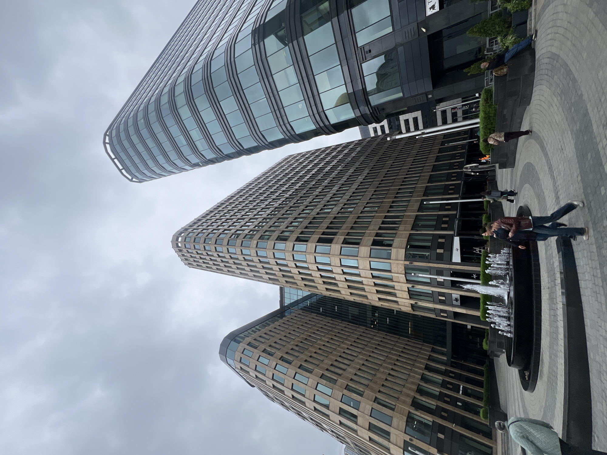 The photograph depicts a modern urban landscape, likely located in Moscow, Russia, based on the architectural style. Three high-rise buildings dominate the frame, each displaying a unique design. The topmost building has a curved, glass facade, while the middle one exhibits a grid-like pattern of windows with a light-brown exterior. The bottom building has a more rounded design with a similar grid pattern.
At ground level, there's a circular area with a fountain in the center, surrounded by a paved surface. Several individuals are visible, some of whom appear to be interacting with the fountain area. Green foliage is interspersed among the buildings, adding a touch of nature to the scene. The sky overhead is overcast, providing soft, diffused lighting.