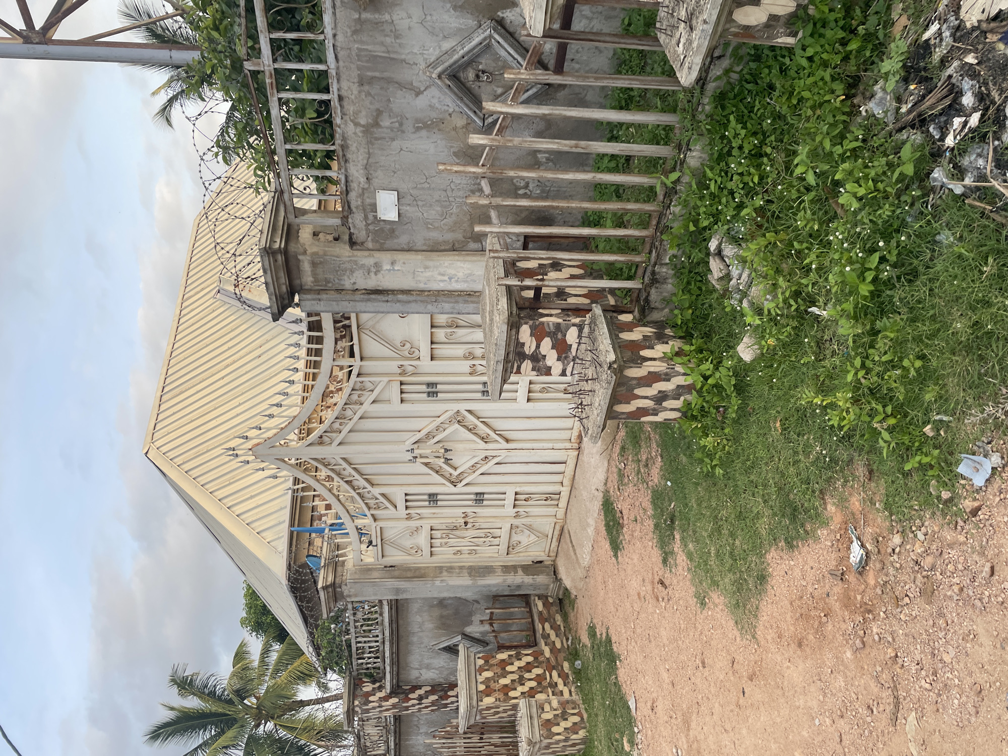 The image shows the exterior of a property in Jalingo, Nigeria. A large, ornate white metal gate is the central feature, flanked by concrete pillars decorated with patterned tiles. To the left of the gate, a concrete wall with similar tile accents encloses the property, and a tall palm tree is visible behind it. A barbed wire fence runs along the top of the wall. To the right of the gate, a low wooden fence with vertical slats separates the property from a patch of overgrown grass and weeds, with some debris visible. The ground in front of the gate is a mix of dirt and gravel. The sky is overcast.