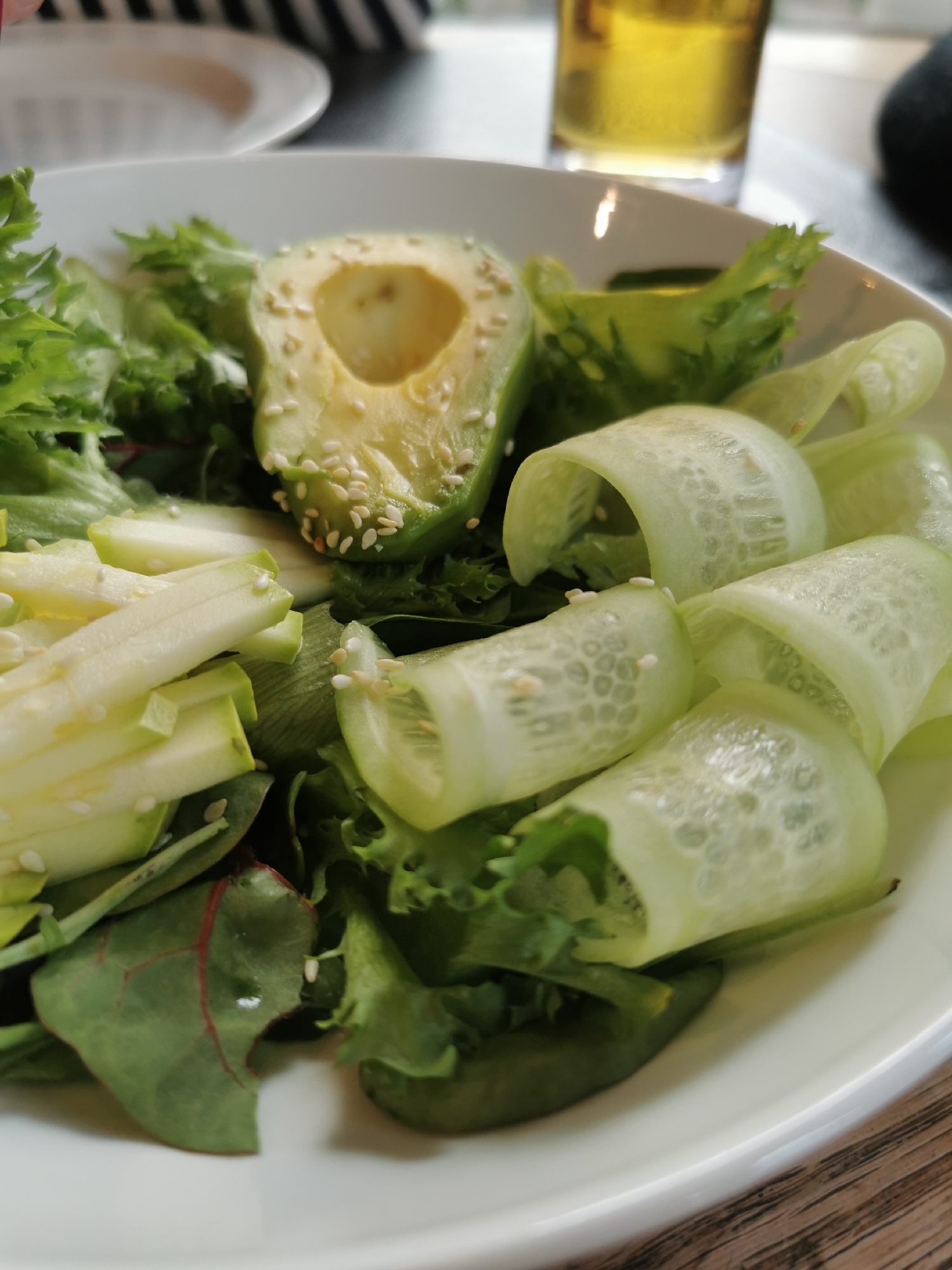 A close-up shot focuses on a white ceramic bowl filled with a variety of fresh salad ingredients. The salad features leafy greens, a halved avocado with sesame seeds sprinkled on its surface, thinly sliced cucumbers rolled into decorative shapes, and pale green zucchini strips. A tall glass of amber-colored liquid, likely beer, sits partially visible in the upper right of the frame, suggesting a meal setting. In the background, a black and white striped object and another white plate are discernible but out of focus.
Given the presence of Eastern European cuisine elements like beer and fresh salad, it is possible the scene is located in the city of Moscow, Russia.