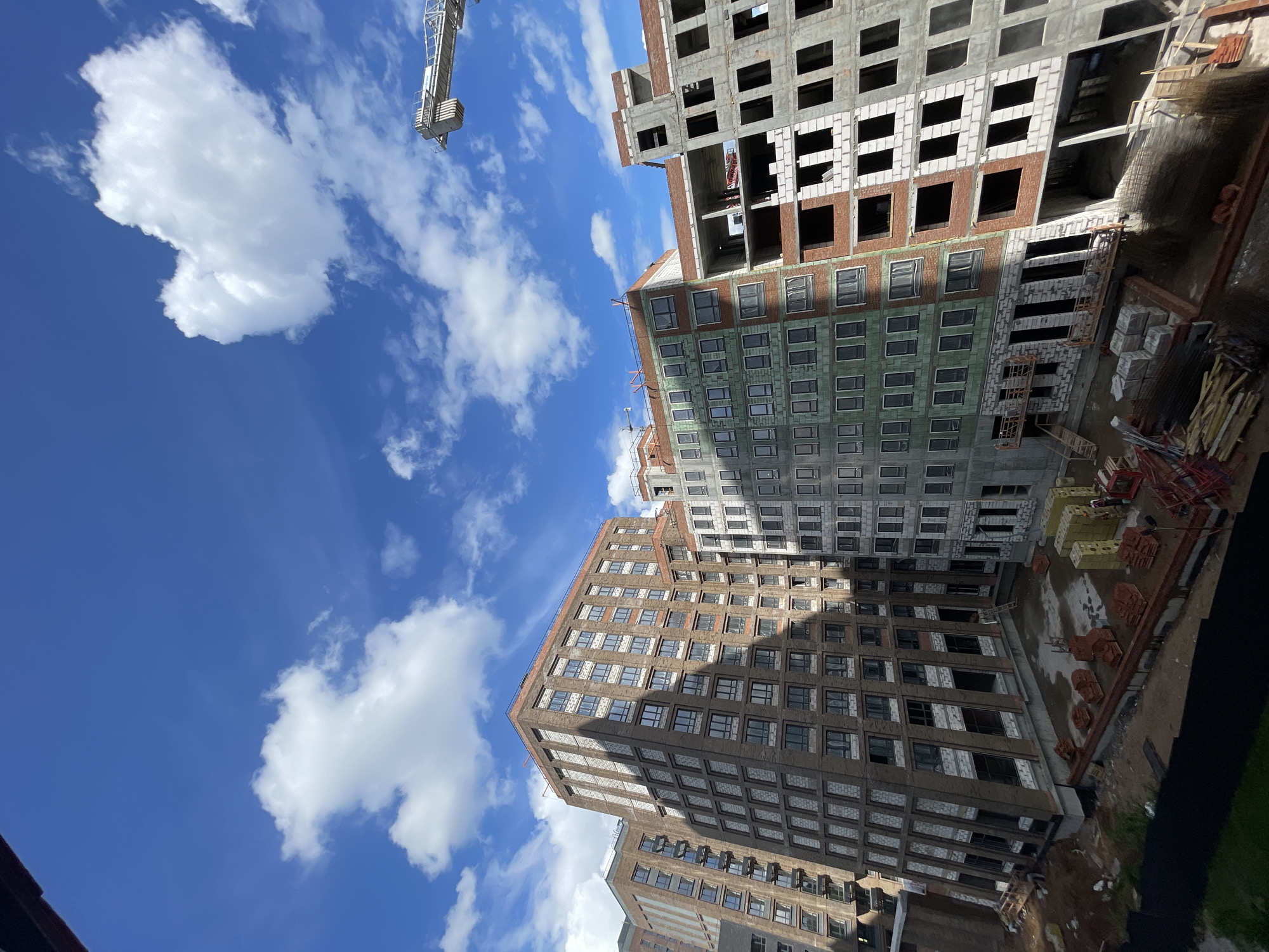 The image shows a construction site in an urban area. Several multi-story buildings are under construction, with various stages of completion evident. The buildings have a rectangular shape and consist of multiple floors. Construction materials and equipment are visible, suggesting ongoing construction activities. A crane is present in the background, indicating its use in the construction process. The sky is blue with scattered white clouds, providing a bright backdrop to the scene. The location is in the city of Unknown, Unknown.