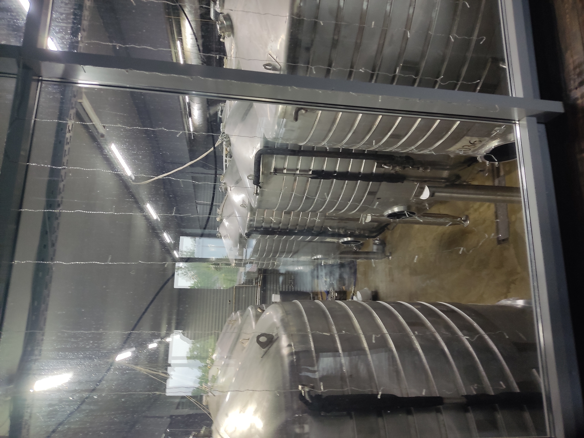 The image shows a view through a window reflecting both the interior and exterior environments. Inside, a series of large, horizontal, cylindrical tanks, likely made of stainless steel, are visible. They are oriented lengthwise, suggesting an industrial setting, possibly a brewery or a chemical processing plant. The tanks are reflective, adding to the visual complexity.
Outside the window, a blurred view of greenery and buildings can be seen.
There is no indication that the location is in Pskov, Russia.