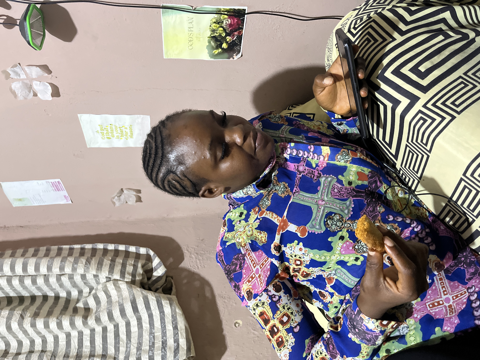 A young Nigerian woman with braided hair is sitting on a bed in a room in Rafin Par, Nigeria. She is wearing a blue and pink patterned shirt and a yellow and black patterned skirt. She is holding a piece of fried food in her left hand and a smartphone in her right hand. The room has a beige-colored wall with several framed pictures and a light fixture hanging from the ceiling. A striped curtain is visible in the background. The scene appears to be an indoor shot taken during the day.