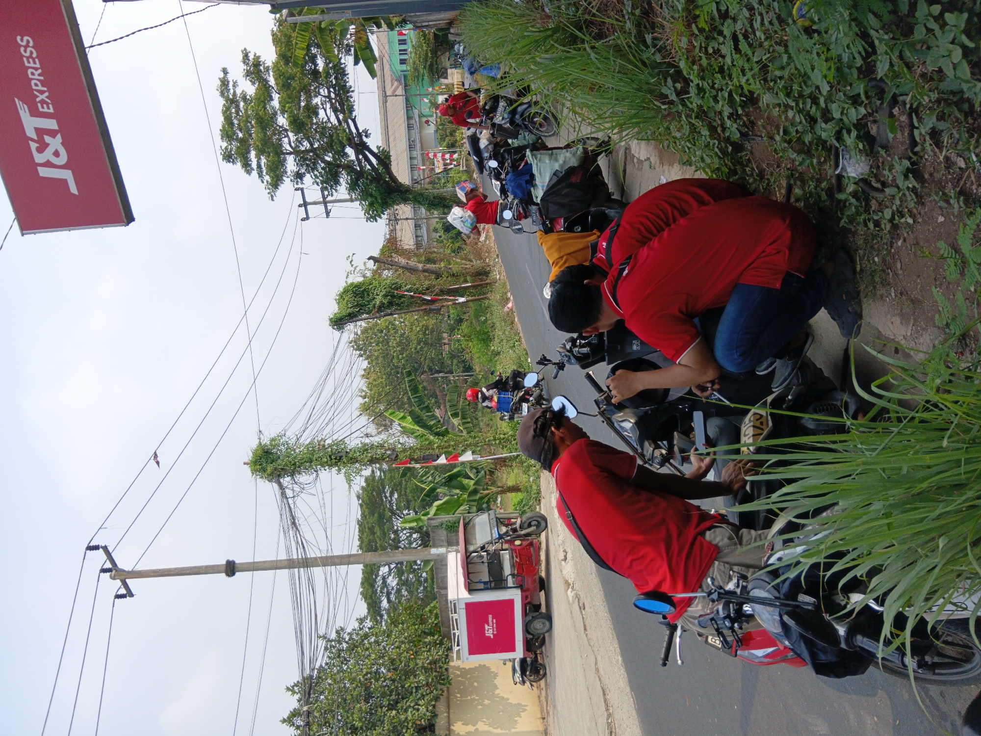 The image depicts a street scene in Bekasi, Indonesia, likely near a J&T Express outpost, given the prominent signage. The foreground features several individuals, primarily wearing red t-shirts, interacting with motorcycles. Two men are crouched or kneeling near motorcycles, one in a red t-shirt and a dark cap, the other in a red t-shirt and blue jeans. A red J&T Express delivery cart is visible parked on the side of the road to the left, with another individual in a red shirt standing near it. Further down the road, another person in a red shirt on a motorcycle, wearing a red helmet, is visible. The environment includes a paved road, a sidewalk with overgrown grass and vegetation, and electrical poles with numerous wires overhead. Buildings are visible in the background, including what appears to be a commercial or industrial facility. The overall impression is one of an active delivery hub or a street corner where logistics operations are taking place.
