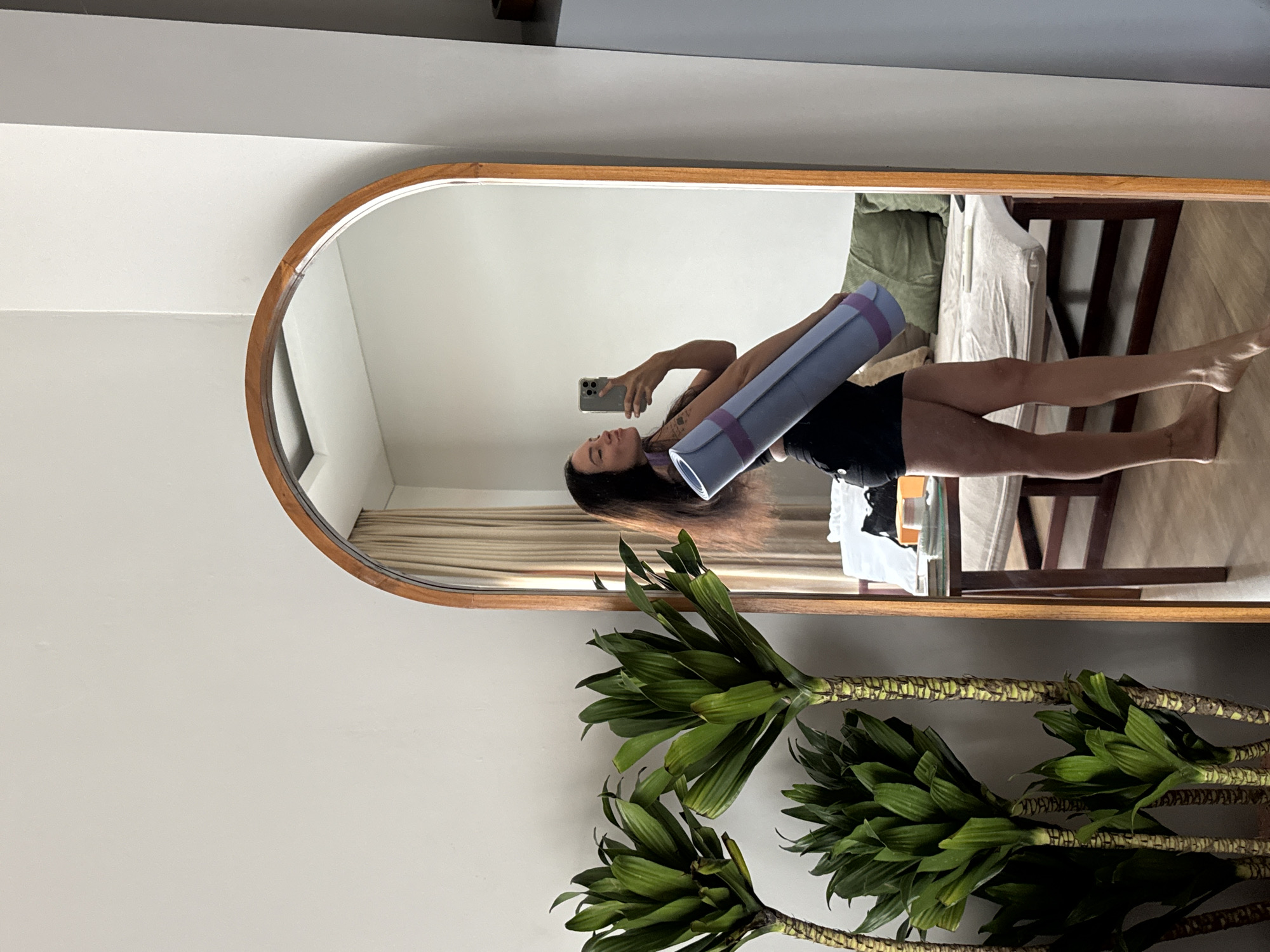 A young woman with dark hair stands in front of a large, arched mirror in what appears to be an indoor setting in North Kuta, Indonesia. She is holding a rolled-up, light purple yoga mat with a purple strap over her right shoulder. She is wearing black shorts and a dark top, and her left arm is extended, holding a smartphone up to take a selfie. Her mouth is puckered as if kissing the camera. The mirror reflects the room behind her, which includes a wooden table with some items on it, a couch with green pillows, and a window with beige curtains. In the foreground, to the left of the mirror, are three large potted plants with broad green leaves. The floor is wooden, and the wall is painted a light color.