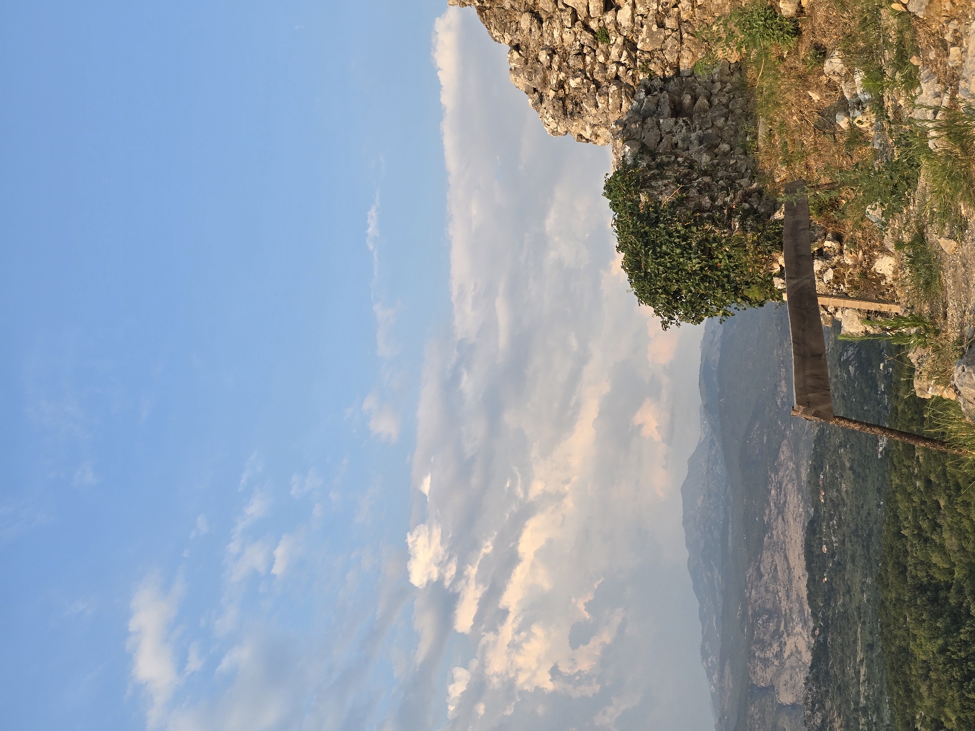 The image depicts a view from the fortress of Medun, Montenegro. In the foreground, a section of ancient stone wall is visible on the right, overgrown with a cluster of green ivy on the left. A wooden plank is positioned horizontally across the scene, supported by a rustic pole. The ground is uneven, covered with rocks, dry grass, and sparse green vegetation. In the middle ground, a lush green valley stretches out, dotted with scattered houses. The background features a range of hazy, majestic mountains under a vast blue sky filled with scattered white and pinkish clouds, illuminated by the warm glow of the setting sun.