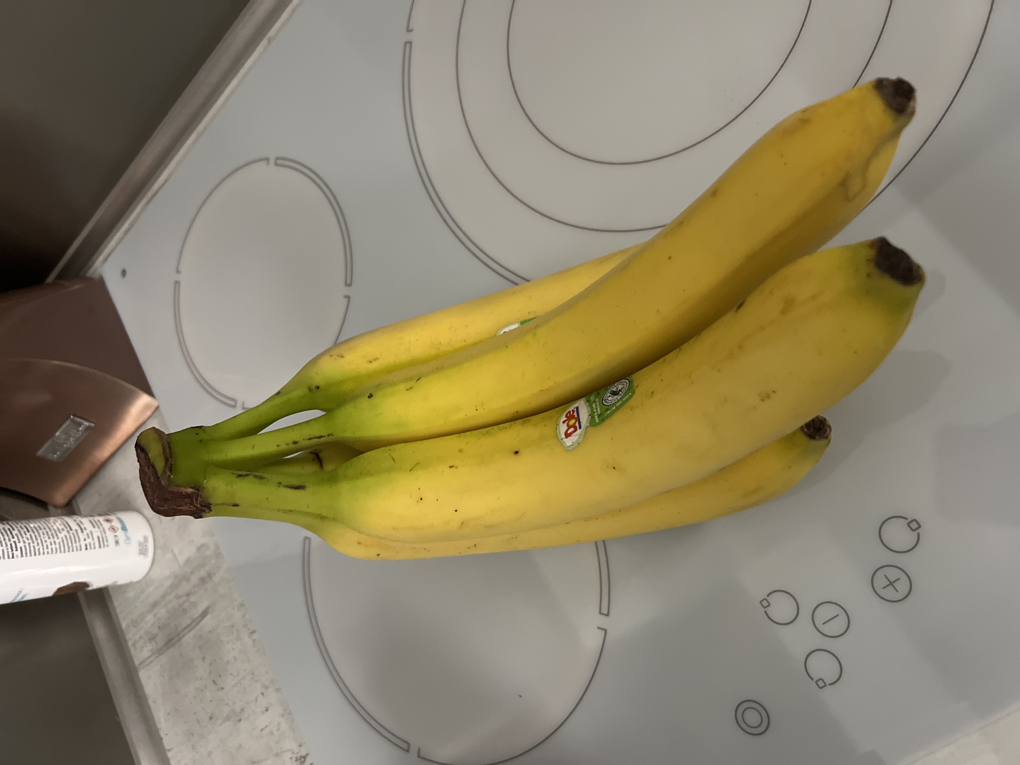 A bunch of bananas sits on a white stovetop with circular burner markings and touch controls. The bananas are mostly yellow with some green near the stems, and a Dole brand sticker is visible on one of them. A partially visible container and a small, metallic brown object are to the left of the banana stems.
The location cannot be determined from the image. There is no information on city or country in the image.
