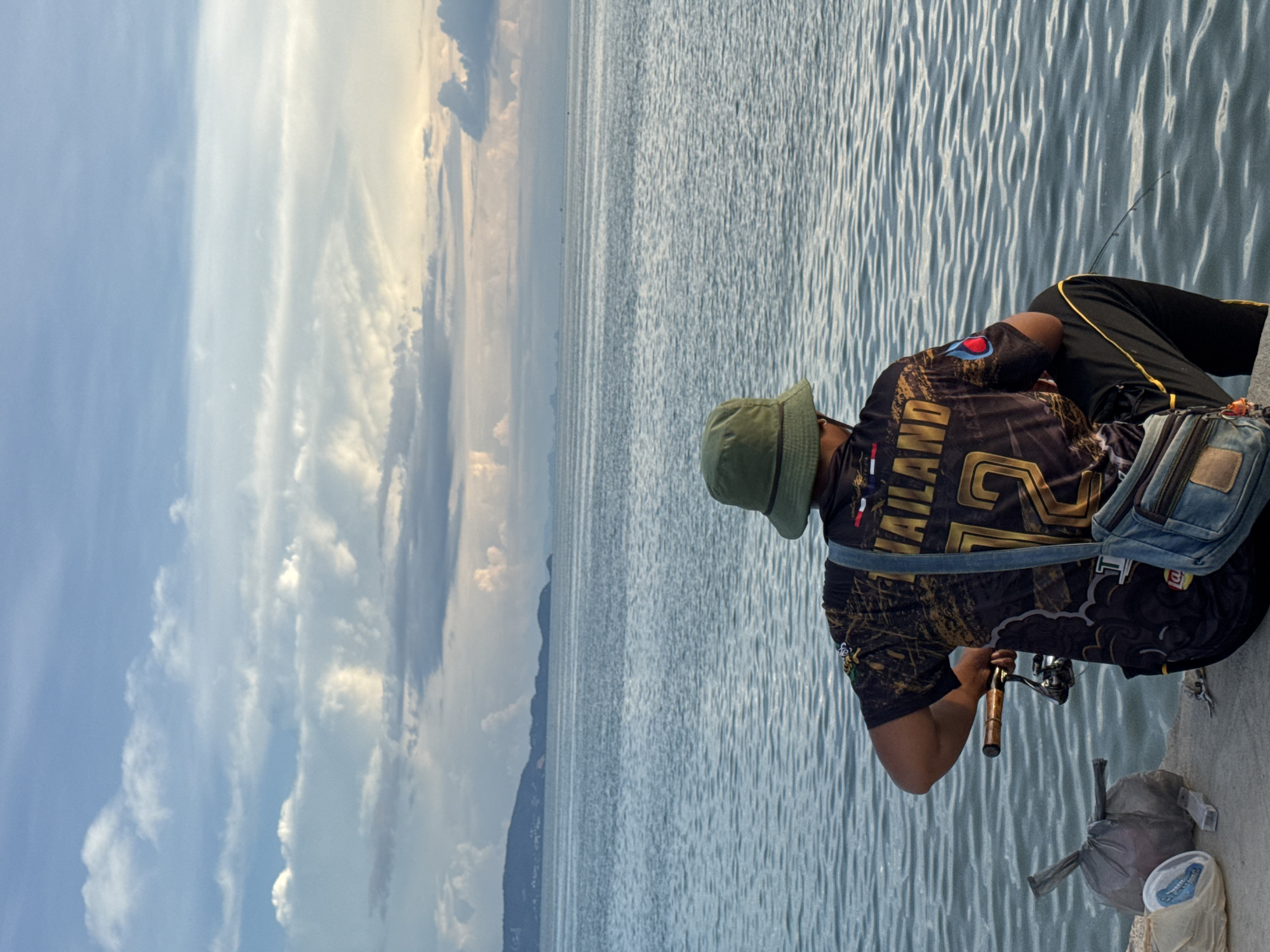 An individual is fishing from a concrete pier at the sea in ตำบลเกาะพะงัน, Thailand. The person is wearing a green bucket hat, a black t-shirt with gold lettering and designs, black pants with a yellow stripe, and a blue denim cross-body bag. They are holding a fishing rod, with the tip extending into the water. Next to them on the pier are two plastic bags, one grey and one white. The sea is calm with small ripples, and the sky is filled with clouds, some of which are illuminated by the sun. In the distance, a hazy landmass with hills can be seen on the horizon.