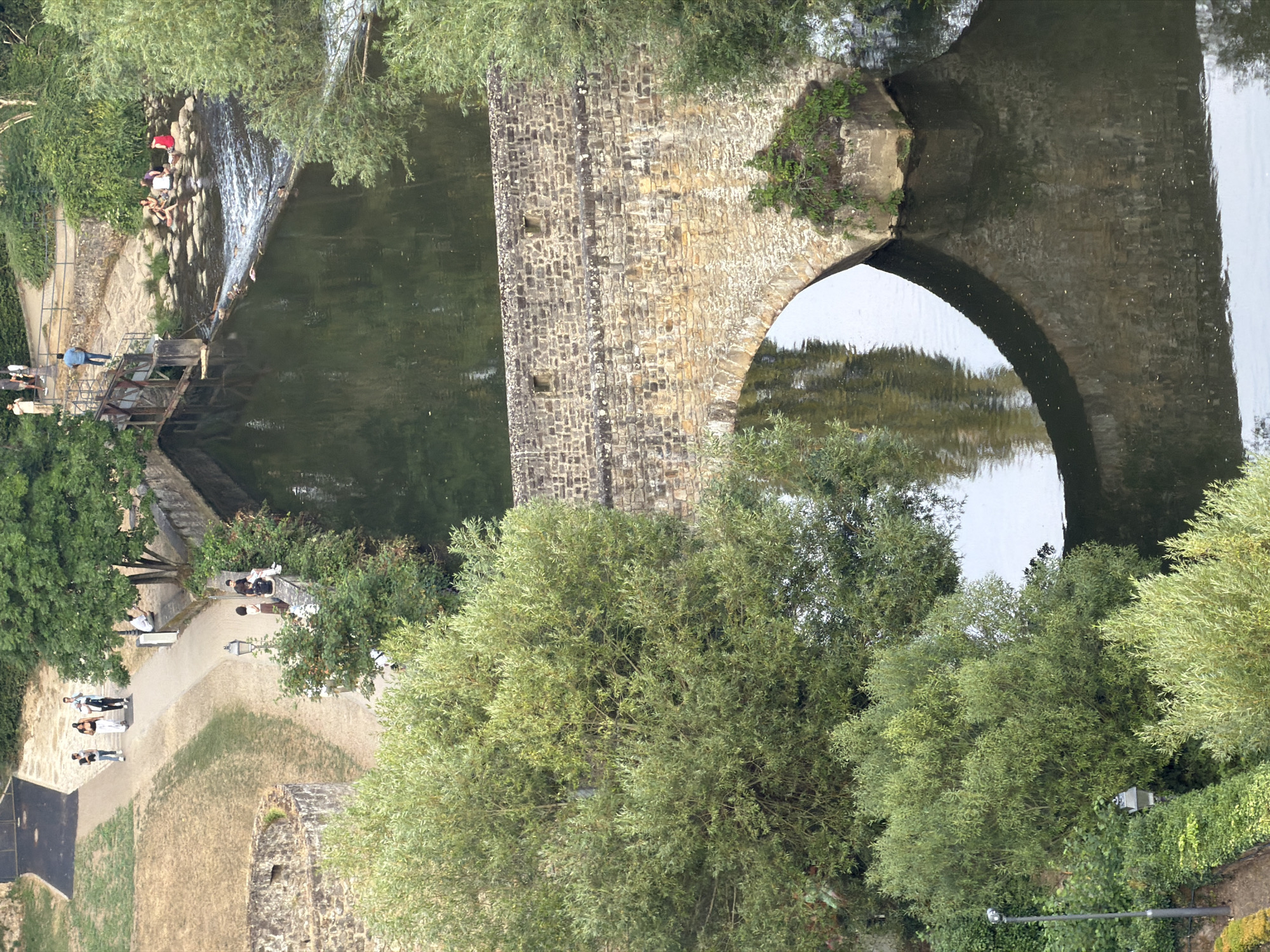 This is an aerial view of a stone bridge arching over a body of water, likely the Alzette River, in Luxembourg City. The bridge is constructed of rough-hewn stones and is surrounded by lush green trees. On the left side of the image, a pathway runs alongside a dry, grassy embankment and a section of stone wall. Further up this pathway, several individuals are seen walking. On the right side of the bridge, the water cascades over a series of rocks, creating a small waterfall. Several people are sitting on the rocks in the river. The water reflects the bridge and the surrounding trees, creating a mirrored image below the arch.