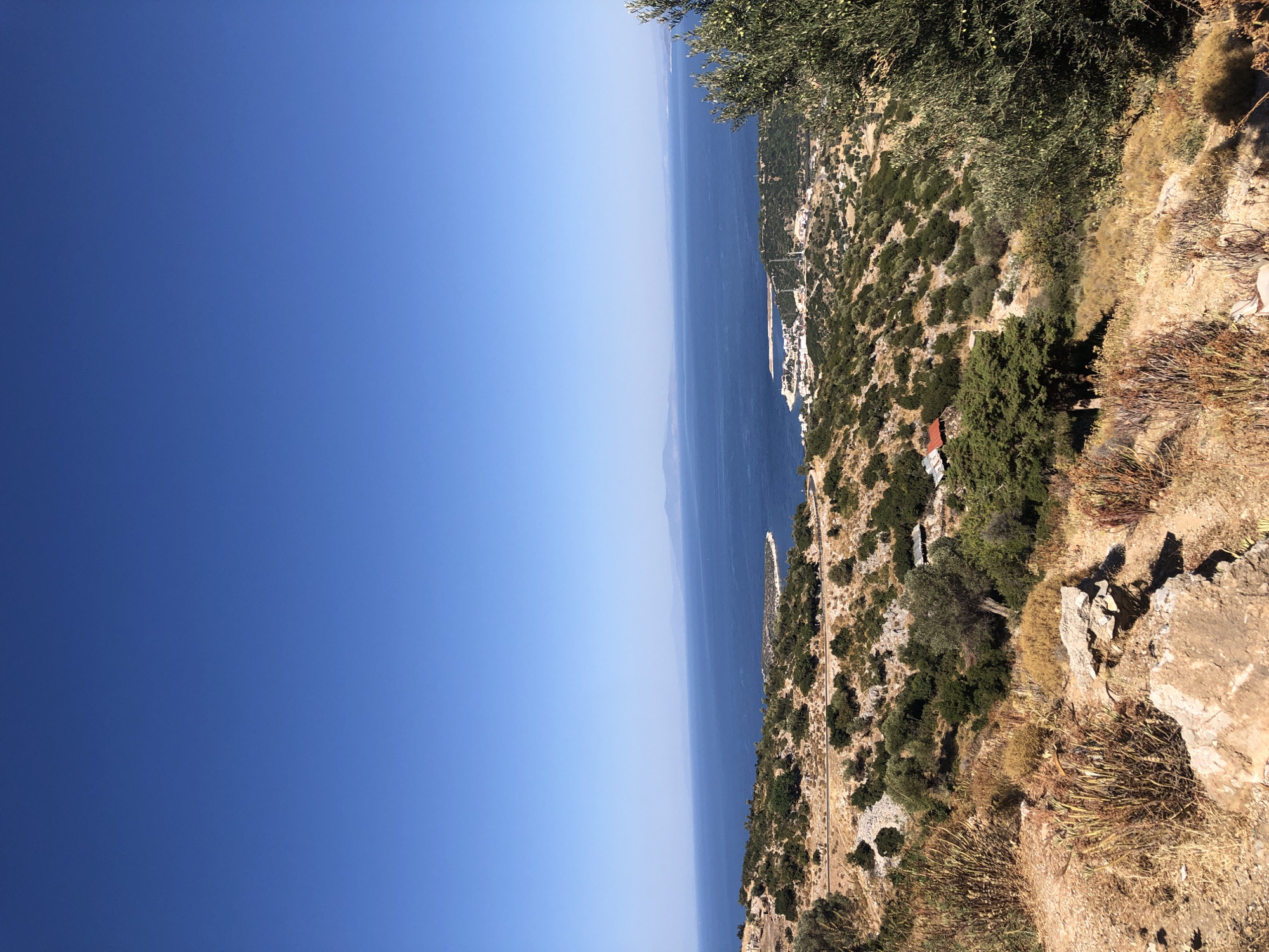 This is a high-angle, wide shot photograph taken on a sunny day overlooking a bay in Karaburun, Turkey. The foreground is dominated by dry, rocky terrain with sparse, sun-bleached vegetation and scattered rocks. A winding road is visible on the left side of the frame, leading down towards the bay.
In the mid-ground, the landscape transitions to a hillside covered in dense green shrubs and scattered olive trees. A few small buildings with red roofs are nestled amongst the foliage. The bay itself is a vibrant blue, with a small harbor area visible in the distance. A cluster of white buildings forms a town along the coast, with a breakwater extending into the water.
The background features the vast expanse of the sea, stretching out to a hazy horizon where distant, muted blue landmasses are visible. The sky is a clear, deep blue, indicating fair weather conditions. The overall impression is one of a dry, Mediterranean coastal landscape with a town nestled by the sea.