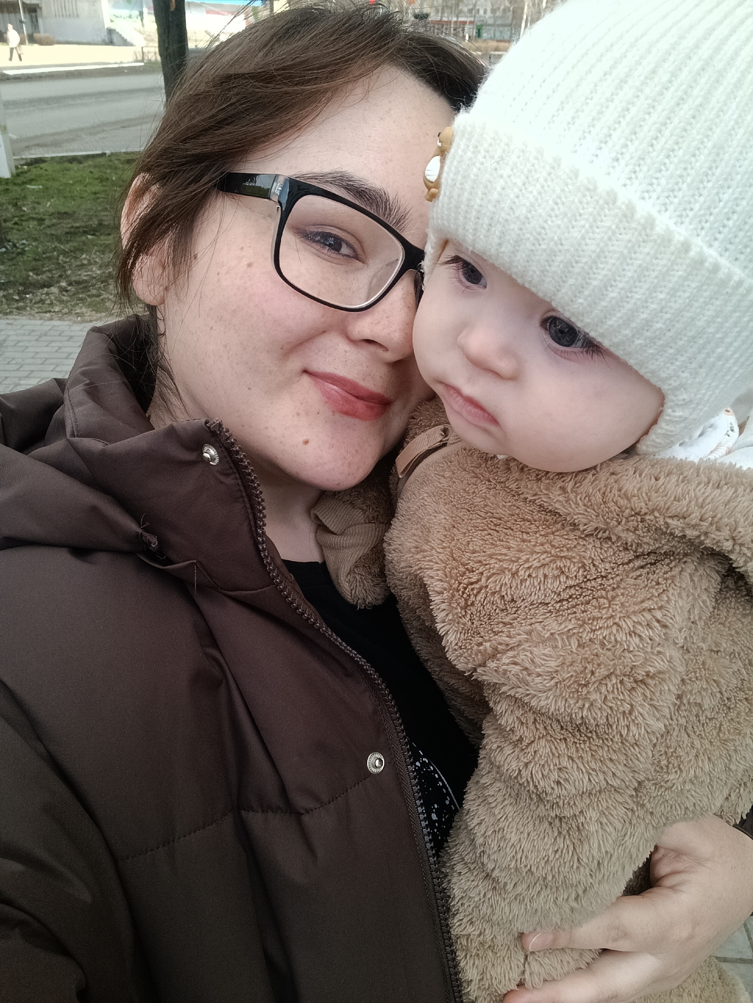A close-up outdoor portrait shows a woman holding a baby in Chaikovsky, Russia. The woman, positioned on the left, has dark hair and wears black-framed glasses, subtly smiling at the camera. She has visible freckles and wears a dark brown puffer jacket over a dark top. Her lips have a reddish-brown tint.
The baby, held close by the woman on the right, wears a thick, white knitted beanie and a cozy, light brown, fuzzy romper or jacket. A small, decorative brown clip, possibly shaped like an animal, is visible near the baby's temple on its hat. The baby has dark eyes and a neutral expression, looking intently towards the right side of the frame.
The background is softly blurred but shows an urban or suburban outdoor setting. A paved surface, possibly a sidewalk or road, is visible, along with patches of green ground cover and distant buildings under bright daylight. The subjects' attire suggests cool weather conditions. No text is visible in the image.