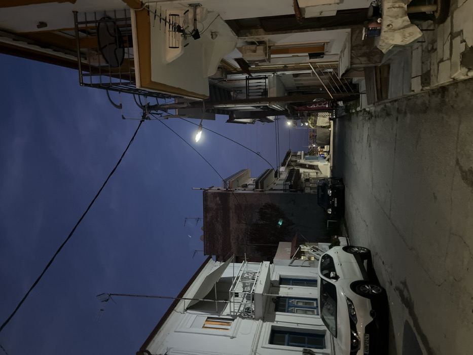 The image shows a narrow street in Limenaria, Greece (latitude: 40.627, longitude: 24.576) at dusk. The street is lined with two-story buildings, most of which are white with balconies. There are a few streetlights illuminating the scene, which is otherwise dimly lit. A white car is parked on the left side of the street, facing the camera. In the distance, there is another car parked on the right side of the street. The sky is a dark blue color, and there are no people visible. The street is paved with asphalt, and there is a sidewalk on the right side. There is a small object on the sidewalk in the bottom right corner of the frame. The buildings on the right side of the frame have an orange and white color scheme and are made of stucco. They have small balconies and a staircase leading to an elevated walkway.