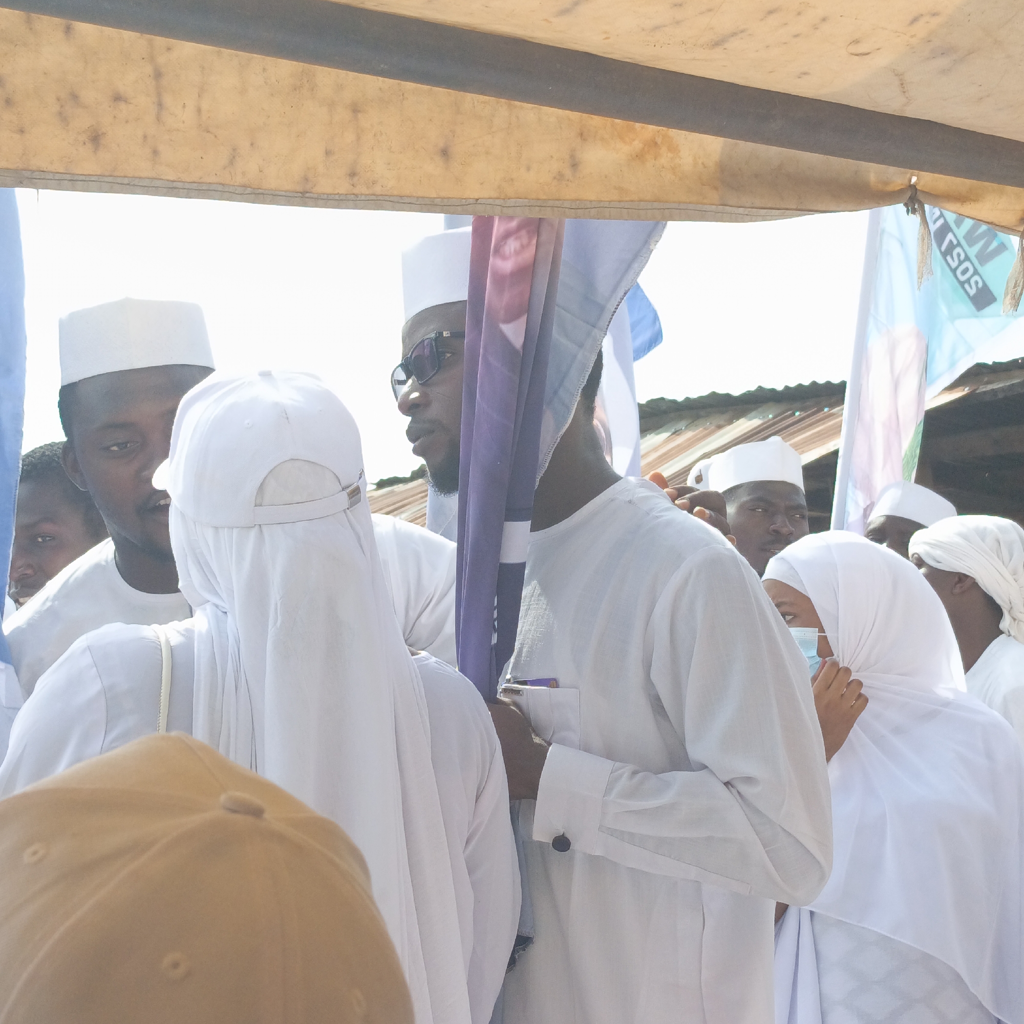 In Akwanga, Nigeria, a group of people wearing white clothing and head coverings are gathered outdoors, under what appears to be a makeshift tent structure. One person is holding a flag with various colors including purple and blue. A man is wearing sunglasses. Several of the women wear white headscarves and one person is wearing a face mask. A tin roof is visible in the background.