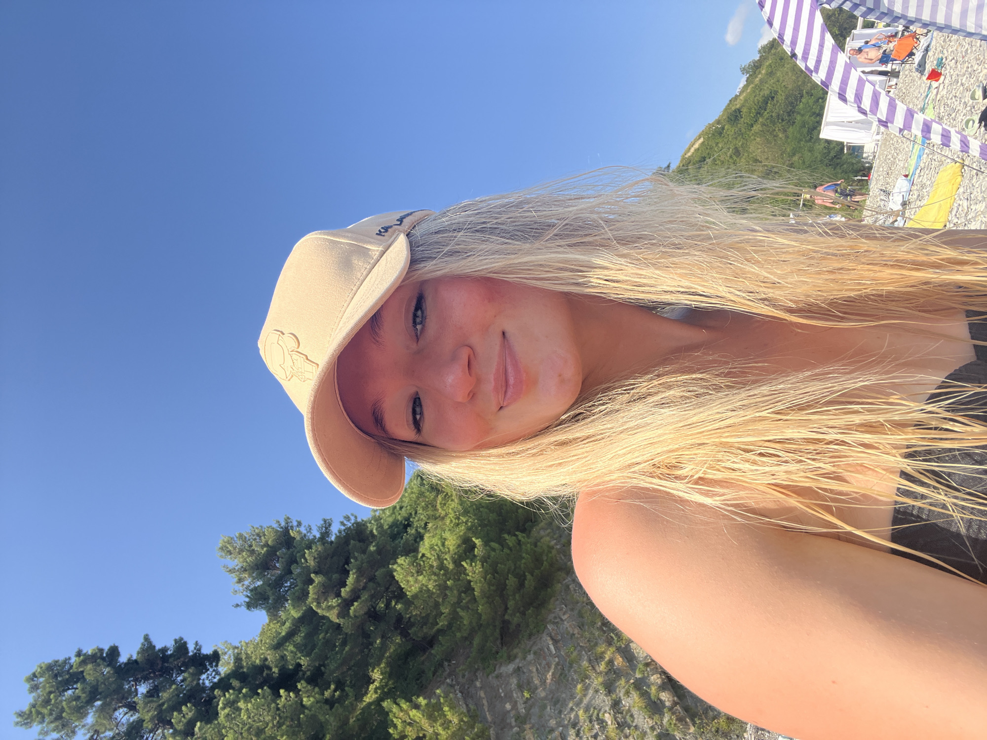 In a sunlit outdoor scene on a rocky beach in Tuapse, Russia, a young Caucasian woman with blonde hair, wearing a tan baseball cap and a dark swimsuit, smiles at the camera. Her hair is wet and hangs loosely around her shoulders. In the background, a steep, tree-covered cliff rises sharply against a clear blue sky. Further down the beach, several other people are visible, some standing and others sitting under beach tents. A purple and white striped tent is prominent in the foreground. The overall impression is of a warm, bright day at the seaside.