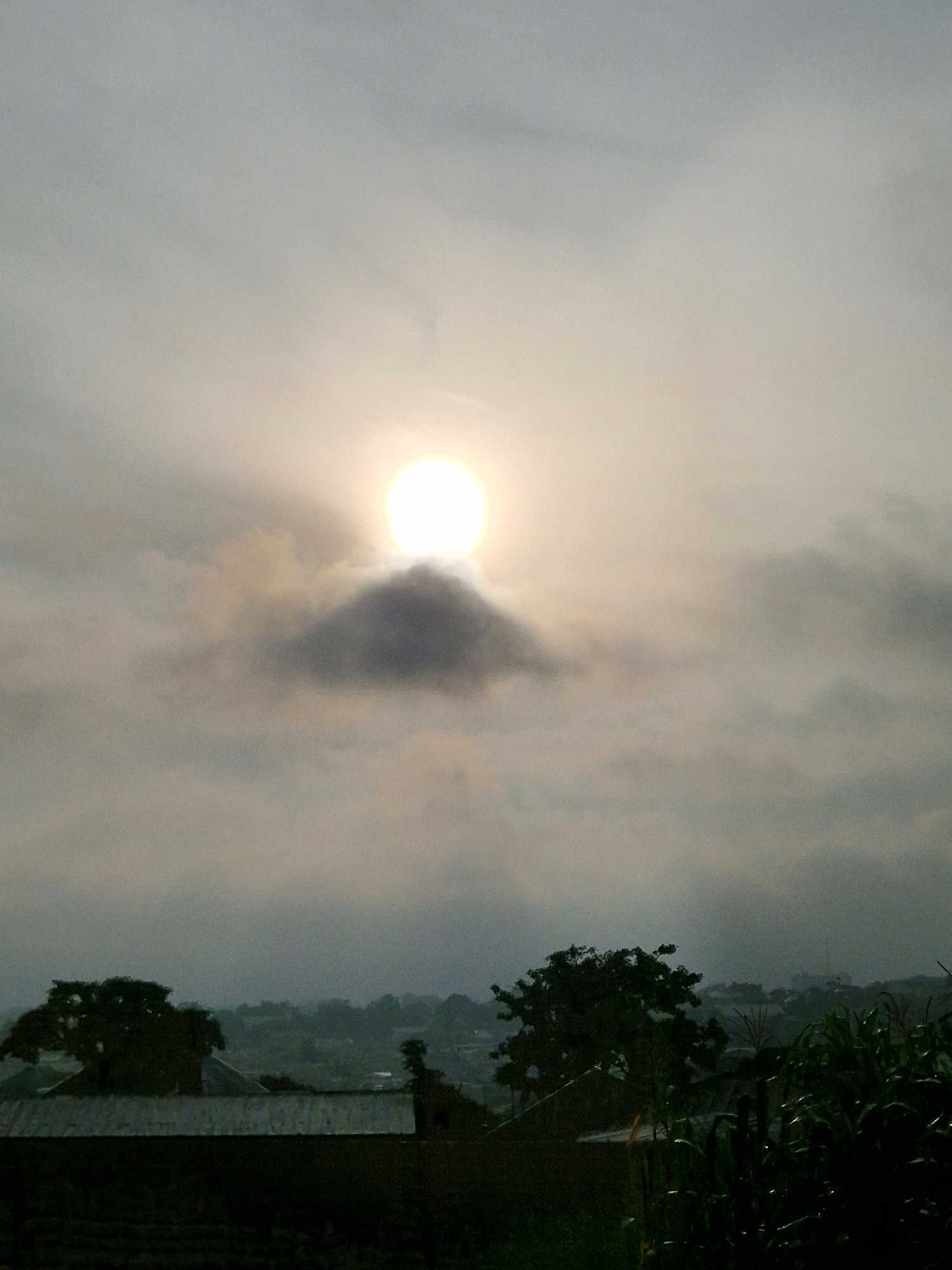 The image depicts a landscape with a hazy sky where the sun is partially obscured by clouds. In the foreground, there are structures that appear to be rooftops of buildings, possibly homes, with some vegetation, including what looks like corn stalks, to the right. In the middle ground, there are trees silhouetted against the sky. The background shows a distant view of a settlement with buildings and more trees, suggesting a rural or semi-urban setting. The overall atmosphere is muted due to the overcast sky and haziness. The image is not specific enough to identify the exact location within Kabong, Nigeria, but it conveys a scene of an overcast day in a populated, vegetated area.