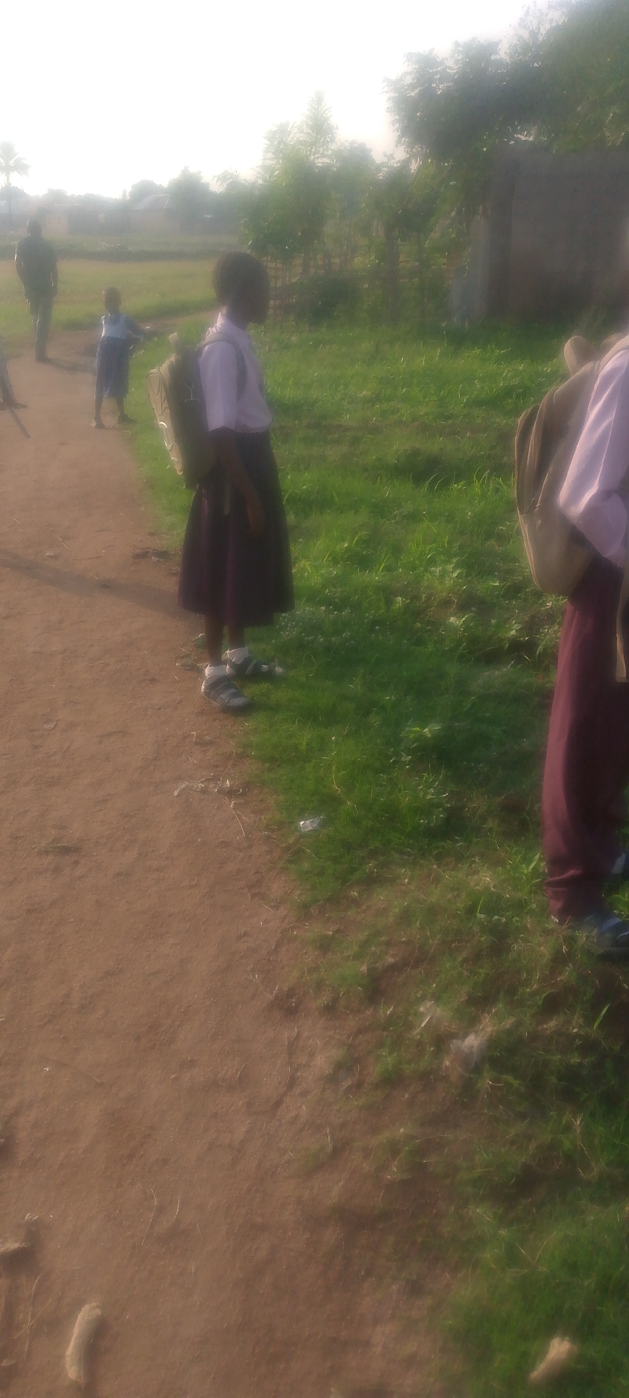 The image depicts a group of children in what appears to be a rural or suburban area, possibly in Jalingo, Nigeria, given the context provided. The scene is outdoors, with a dirt path on the left and a grassy area on the right. In the foreground, a young girl in a school uniform, consisting of a white shirt and a dark skirt, stands with a backpack. Her feet are clad in sandals. Behind her, further down the path, another child in a similar uniform is visible. In the background, a man in dark clothing is walking away from the camera. The environment features sparse vegetation, including trees and grass, and a hazy, bright sky, suggesting either early morning or late afternoon light. The overall impression is that of children on their way to or from school.