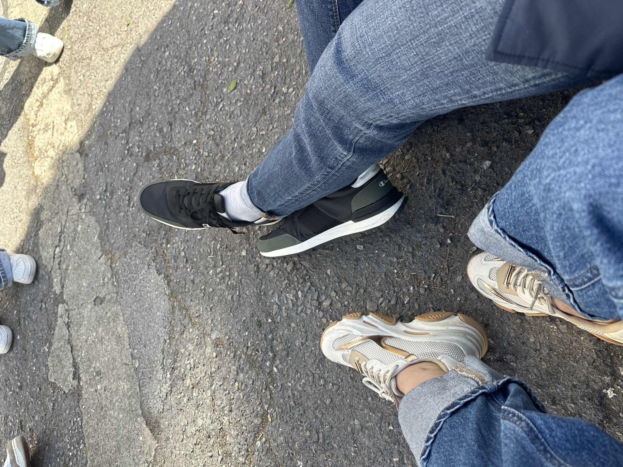 The image shows a section of asphalt pavement with portions of at least three individuals present. All individuals are wearing blue denim jeans. One individual wears black Champion athletic shoes with white socks. Another individual wears beige and white athletic shoes. Portions of other people and their feet, shod in white athletic shoes, are visible along the upper left edge. The asphalt is gray, showing wear and some cracking. The scene appears to be outdoors. The location is unknown and there is no supporting evidence to assume the image was taken in Sofia, Bulgaria.