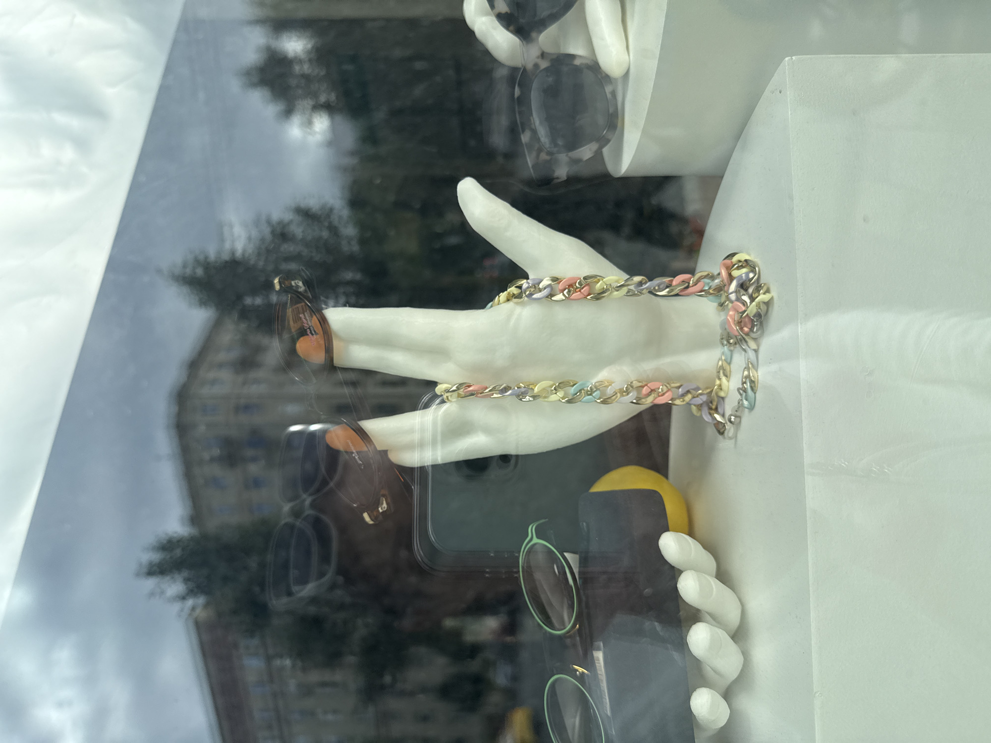 The image shows a display window of a store, possibly an optician or fashion boutique, with sunglasses and a decorative chain. The display features a white mannequin hand wearing a colorful, chunky chain necklace. On the left, another white mannequin hand holds a pair of sunglasses with green frames. Further to the right, another pair of sunglasses with a marbled pattern is displayed. In the background, reflections on the glass show buildings and trees, suggesting an urban environment. The sky appears overcast. The overall setting suggests a street-level display, likely in a commercial area of Moscow, Russia, given the architectural style of the reflected buildings.