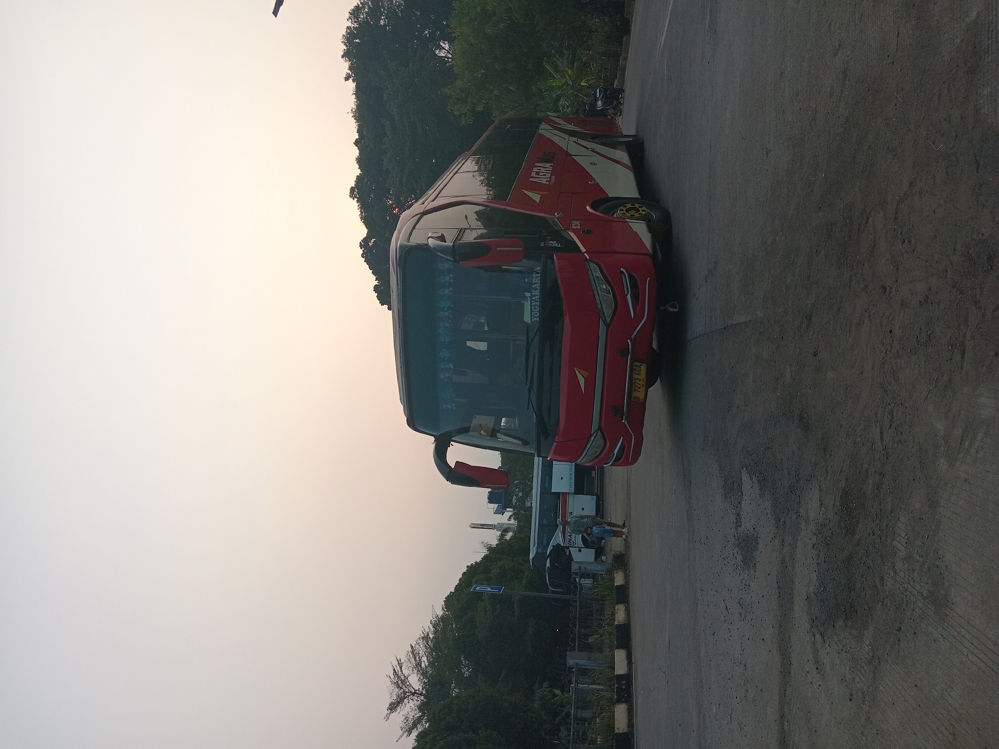 The image shows a red and white bus parked on a paved surface, possibly a bus terminal or parking lot, in Depok, Indonesia. The bus has "AGRA MAS" written on its side, indicating it is part of the Agra Mas bus company. There are other buses visible in the background, along with trees and a utility pole. Two people, a man and a child, are standing near a blue parking sign in the middle ground. The sky is overcast with a soft, diffused light, suggesting it is either early morning or late afternoon. The overall scene is calm and ordinary, depicting a typical day at a transportation hub.