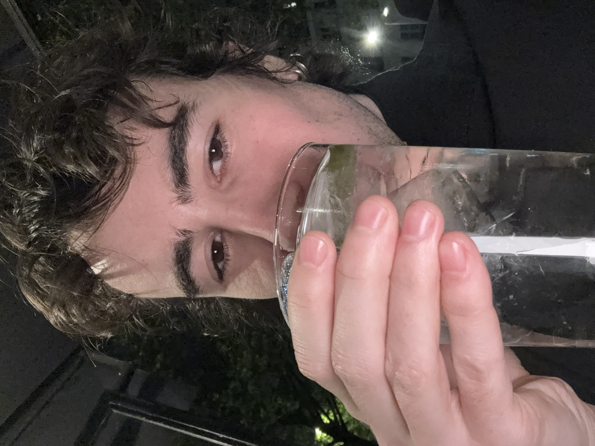 A man with dark curly hair and a small earring is drinking from a clear glass of water. He has brown eyes and thick eyebrows. His hand is holding the glass, and his nails are neatly trimmed. The background shows a dark outdoor setting with some foliage and faint lights, possibly suggesting an evening scene in a restaurant or bar. It is not possible to discern that the location is in Paris, France from the image.