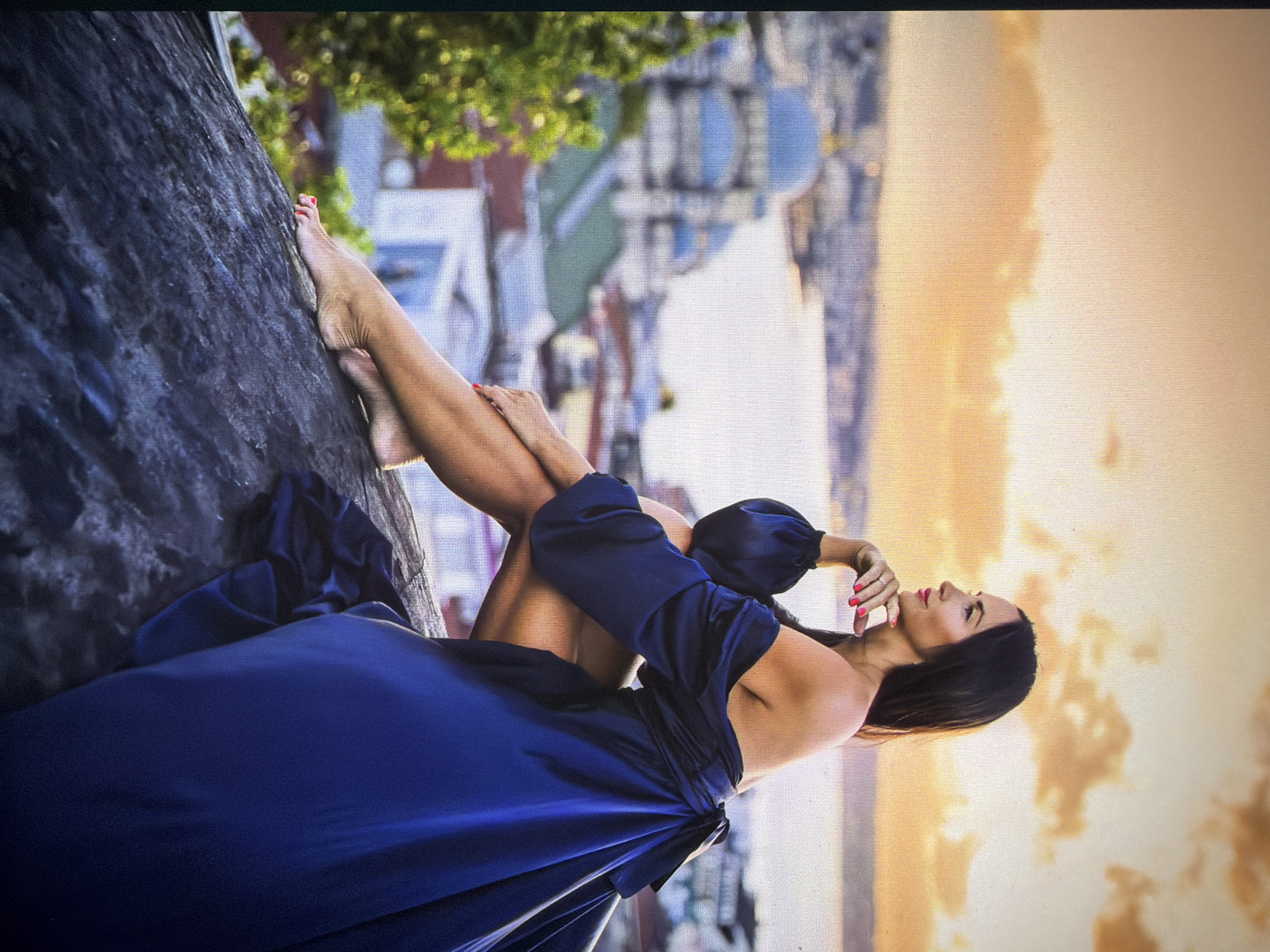 A woman with long dark hair, wearing an off-the-shoulder, flowing navy blue gown, is perched on a rock overlooking a city at sunset. Her legs are crossed, and she is looking to the left, with her chin resting on her hand. The city in the background appears to be a coastal area with domed buildings, possibly the Blue Mosque or Hagia Sophia, suggesting the location is Istanbul, Turkey, not Kadıköy, as Kadıköy is on the Asian side of Istanbul and this view appears to be from the European side looking across the Bosphorus. The sky is a warm gradient of orange and yellow, with scattered clouds, indicating it is either sunrise or sunset. In the foreground, lush green foliage is visible to the left of the woman. The rock she is sitting on is dark and textured.