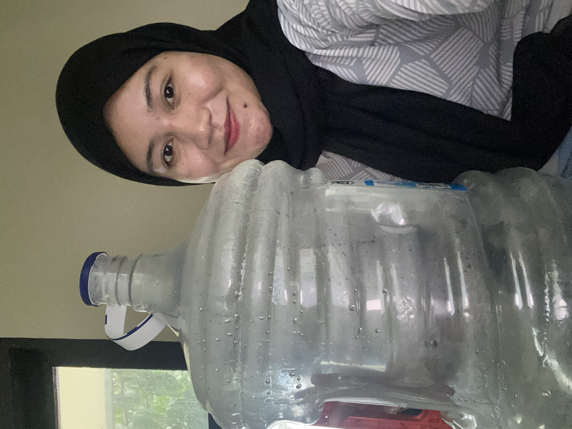 A woman wearing a black hijab and a patterned shirt is posing for a selfie next to a large, clear plastic water jug filled with water. The jug is positioned to the left of the frame and takes up a significant portion of the image. The woman is smiling and her face is well-lit. She is wearing dark lipstick and has a mole on her chin. The background appears to be an indoor setting with a window on the left side, through which greenery can be seen. The overall impression is a casual, everyday moment. The location in Banjarbaru, Indonesia, is an assumption based on the provided context, not explicitly visible in the image itself.