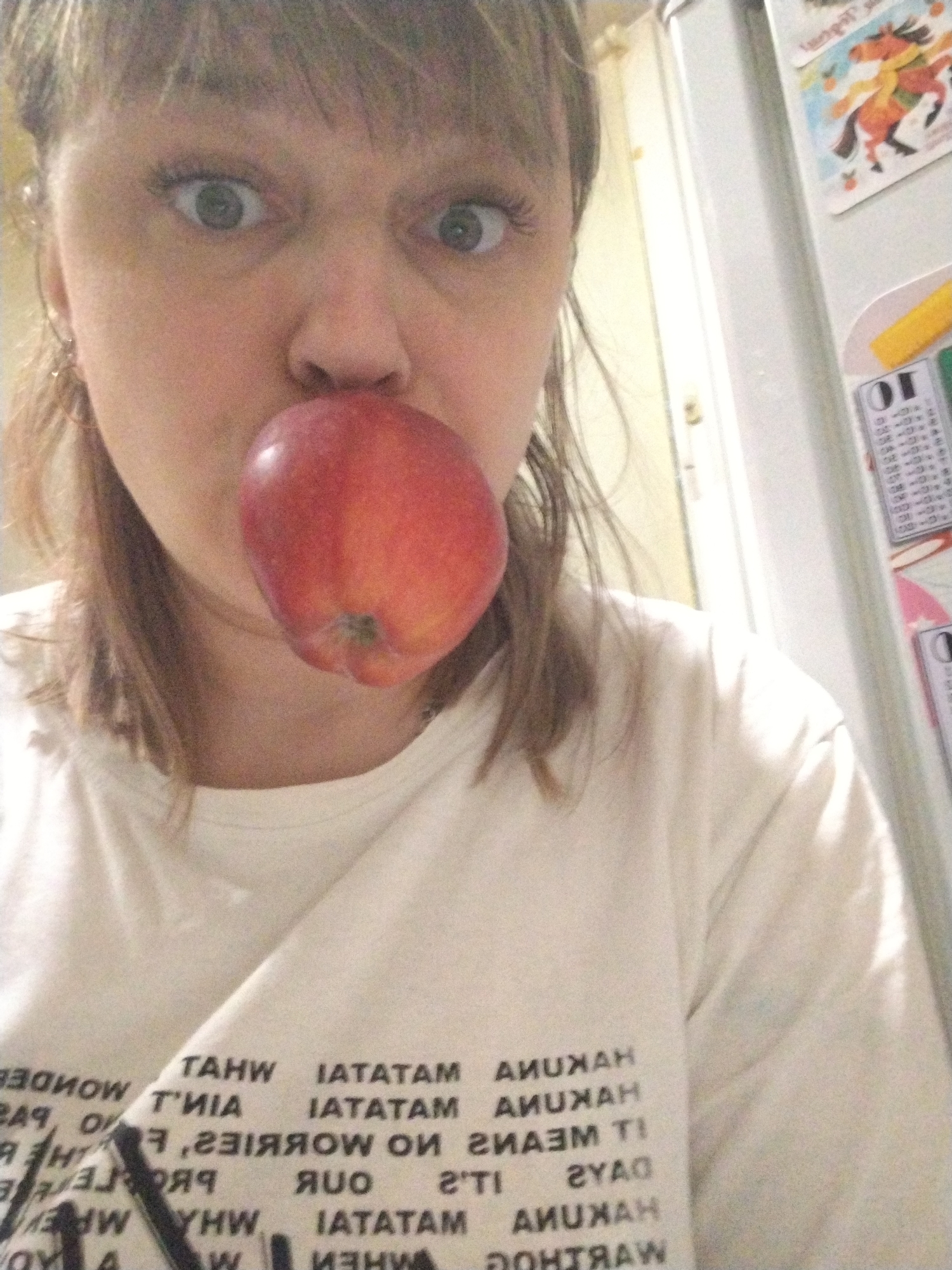A woman is holding a red apple in front of her mouth. She is wearing a white t-shirt with black writing on it. The background includes a refrigerator with magnets and a calendar. The photograph was taken in what appears to be a kitchen. There is no indication this picture was taken in Rusanovo, Russia.