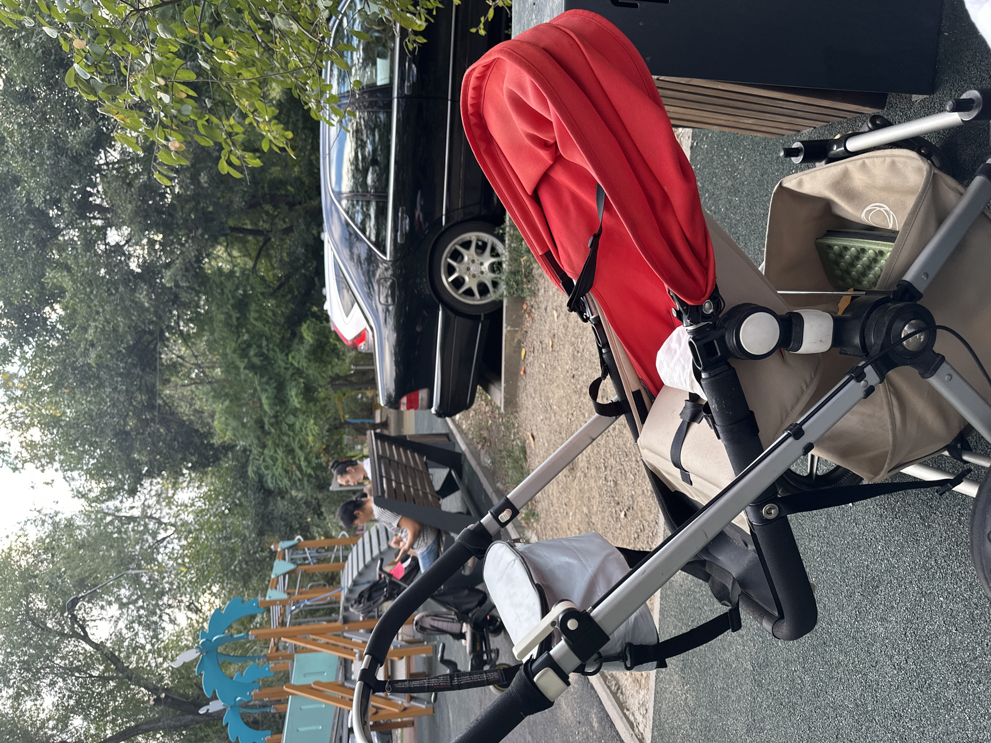 The image depicts an outdoor scene, likely a park or playground, with a focus on a stroller in the foreground. A black luxury sedan is parked in the background, with trees and playground equipment visible. Two women are seated on a bench in the mid-ground, one of whom is looking at her phone. The ground is a textured, dark grey material, possibly rubberized surfacing common in playgrounds. The stroller itself is a prominent subject, featuring a red canopy and a beige fabric seat with straps. A white bag is attached to the stroller's handle. The overall impression is of a moment captured in a public recreational space. The presence of a playground and car suggests an urban environment. Given the context of Almaty, Kazakhstan, this scene is likely taking place in a park within the city.