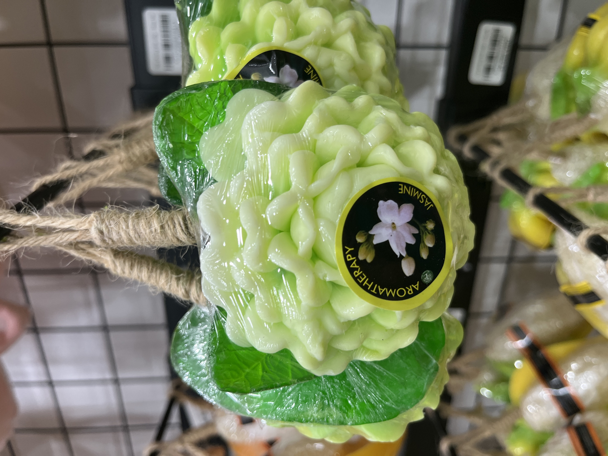 In a retail setting in Nha Trang Ward, Vietnam, a close-up, eye-level shot focuses on three decorative, flower-shaped soaps. These soaps, likely made of a translucent, waxy material, are a pale green and are intricately molded to resemble petals. Each soap is individually wrapped in clear plastic and suspended by a thick, coarse jute rope, which hangs from a dark metal grid.
The soap in the foreground is the most in focus, revealing a circular yellow label adhered to its surface. This label features a black background with white text reading "AROMATHERAPY" in an arc above a delicate illustration of a pale pink jasmine flower with green leaves and buds. The word "JASMINE" is written in white text below the illustration. Another similar soap is partially visible to the right, also bearing a portion of a yellow label.
Behind the main subjects, the background is blurred, showing the dark metal grid and the faint outlines of other merchandise. The lighting appears to be indoor retail lighting, casting some reflections on the plastic wrapping of the soaps. The overall impression is of a display of novelty bath or decorative items in a shop.