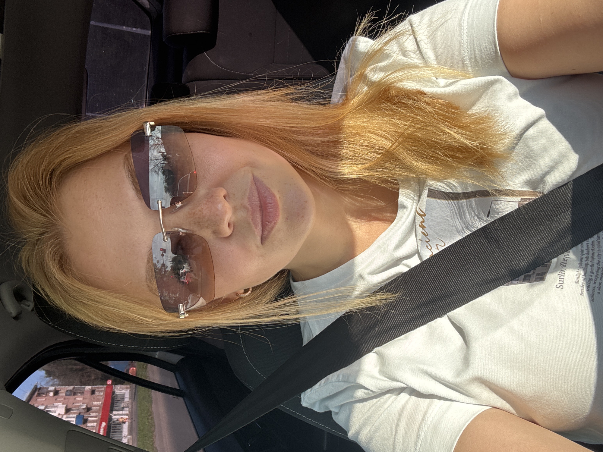 The image shows a young blonde woman with fair skin and freckles, wearing a white t-shirt and sunglasses, sitting in the driver's seat of a car. The seatbelt is visible across her chest. The interior of the car appears to be dark gray. Through the side window, a street scene in Rusanovo, Russia is visible, with a multi-story brick building with red accents, trees, and a traffic light. The woman is looking at the camera, and her hair is a light blonde or strawberry blonde color. Sunlight is casting shadows inside the car.