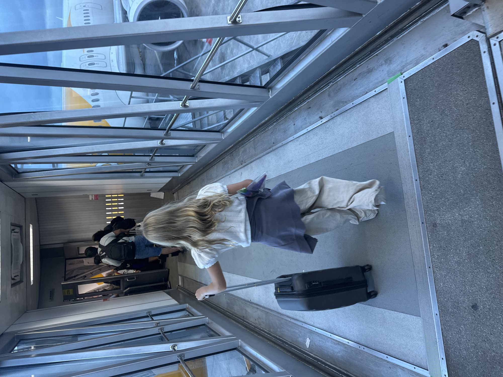 A young girl with blonde hair is walking down an airport jet bridge, pulling a black suitcase. She is wearing a white top and light-colored pants, with a purple garment tied around her waist. Behind her, other passengers are also moving through the jet bridge. The jet bridge has large glass windows on the right, offering a view of an airplane parked outside. The location is identified as Kuta, Indonesia. The environment appears to be an airport terminal, with the jet bridge connecting to the aircraft. The lighting suggests it is daytime, possibly late afternoon or early evening, given the somewhat subdued light. The overall scene captures a moment of travel as people board or disembark from a flight.