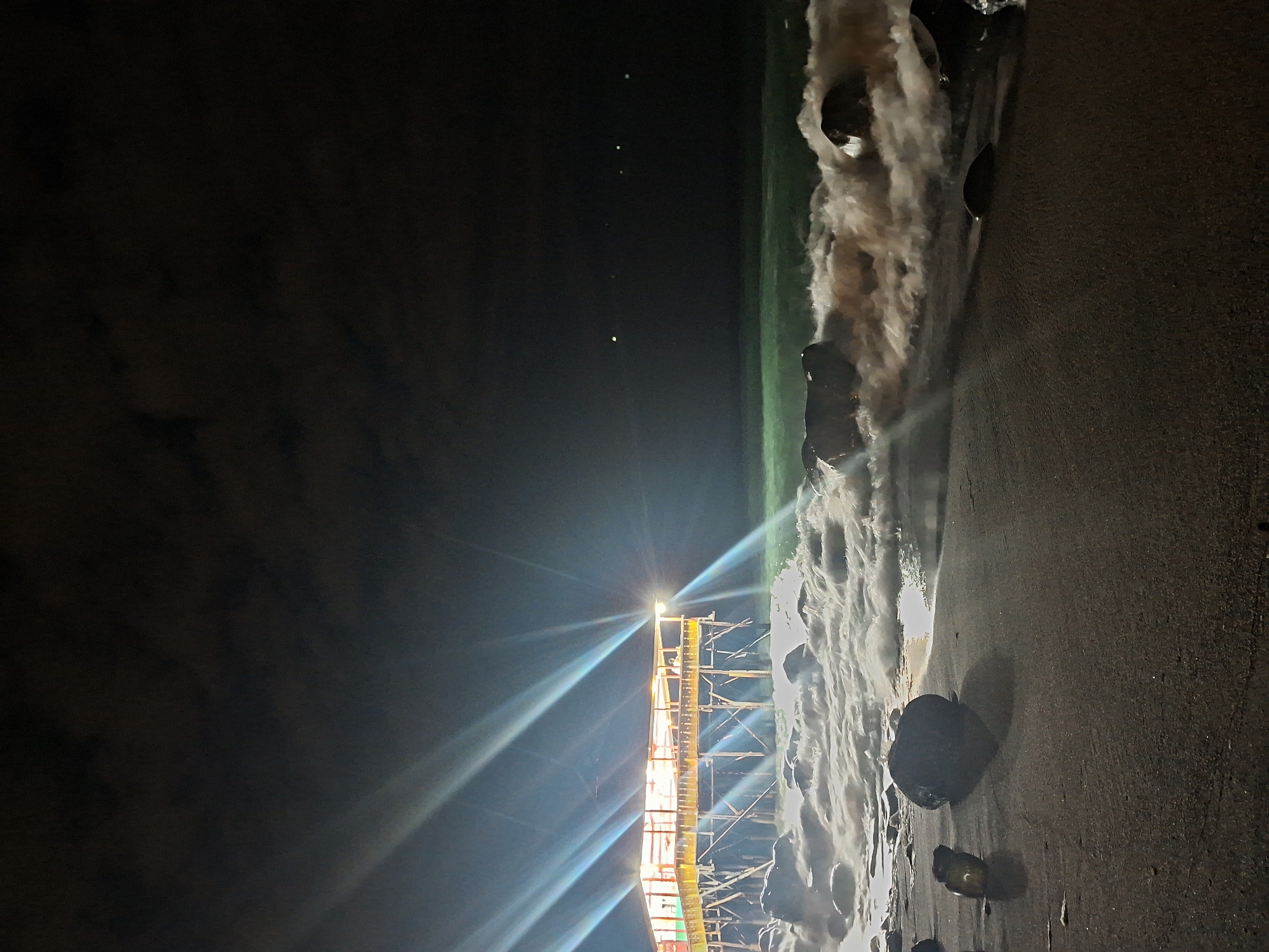 The image depicts a nighttime scene at a beach in Lhokseumawe, Indonesia. Scaffolding with a bright light source is prominently positioned on the beach, casting rays across the scene. Waves crash onto the shore near the scaffolding. Large dark rocks are visible in the sand. The sky is dark with sparse stars. The overall lighting is dramatic due to the bright scaffolding light.