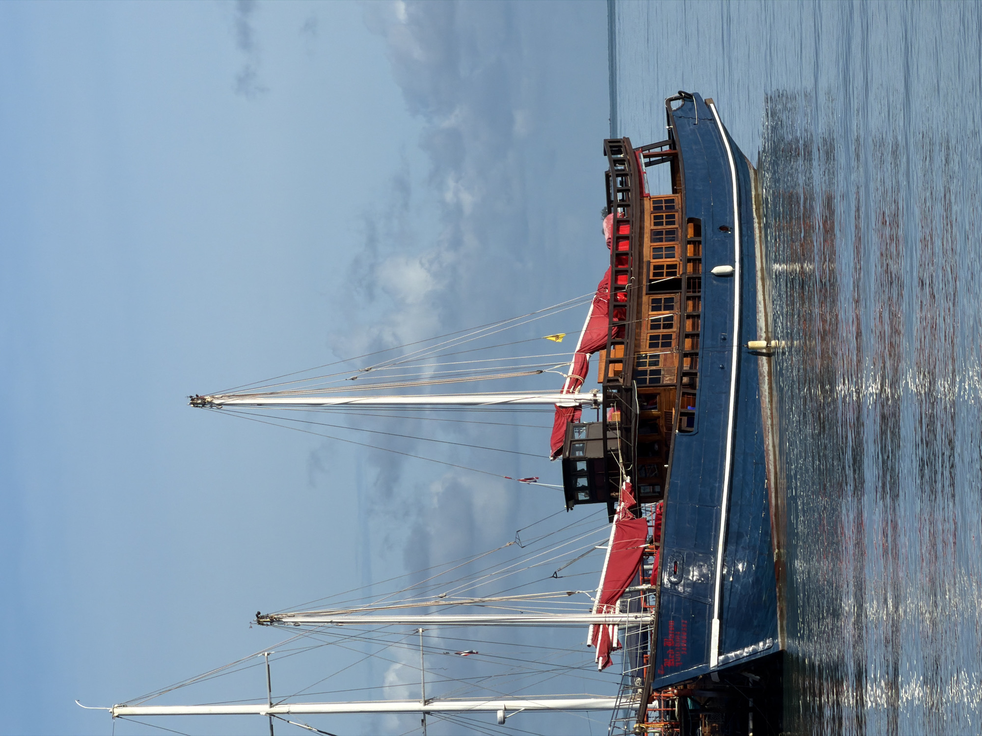 A medium shot captures a large, blue-hulled, wooden-decked boat docked in the clear, calm waters of Baan Bang Rak, Thailand. The boat, featuring multiple masts and red sails, is moored alongside other vessels. The sky above is a mixture of clear blue and scattered clouds. The water's surface reflects the boat and the sky. The scene is still and tranquil.