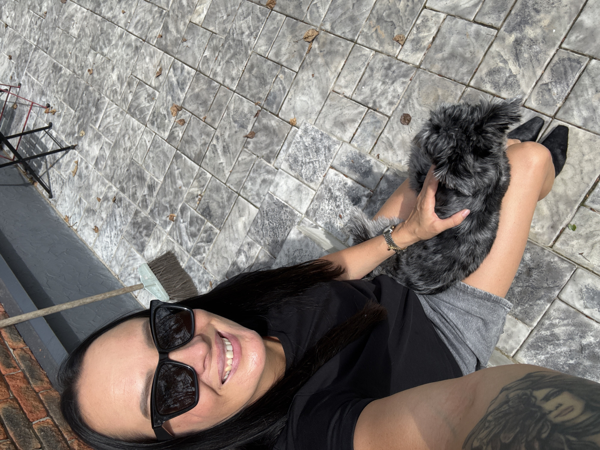 A woman with long dark hair and sunglasses is sitting on a paved patio, holding a small, dark, fluffy dog on her lap. She is wearing a black t-shirt and gray shorts. A broom leans against a brick wall in the background. The patio is made of rectangular gray pavers with some fallen leaves scattered on them. The scene appears to be outdoors on a sunny day. The location is identified as Chaikovsky, Russia.